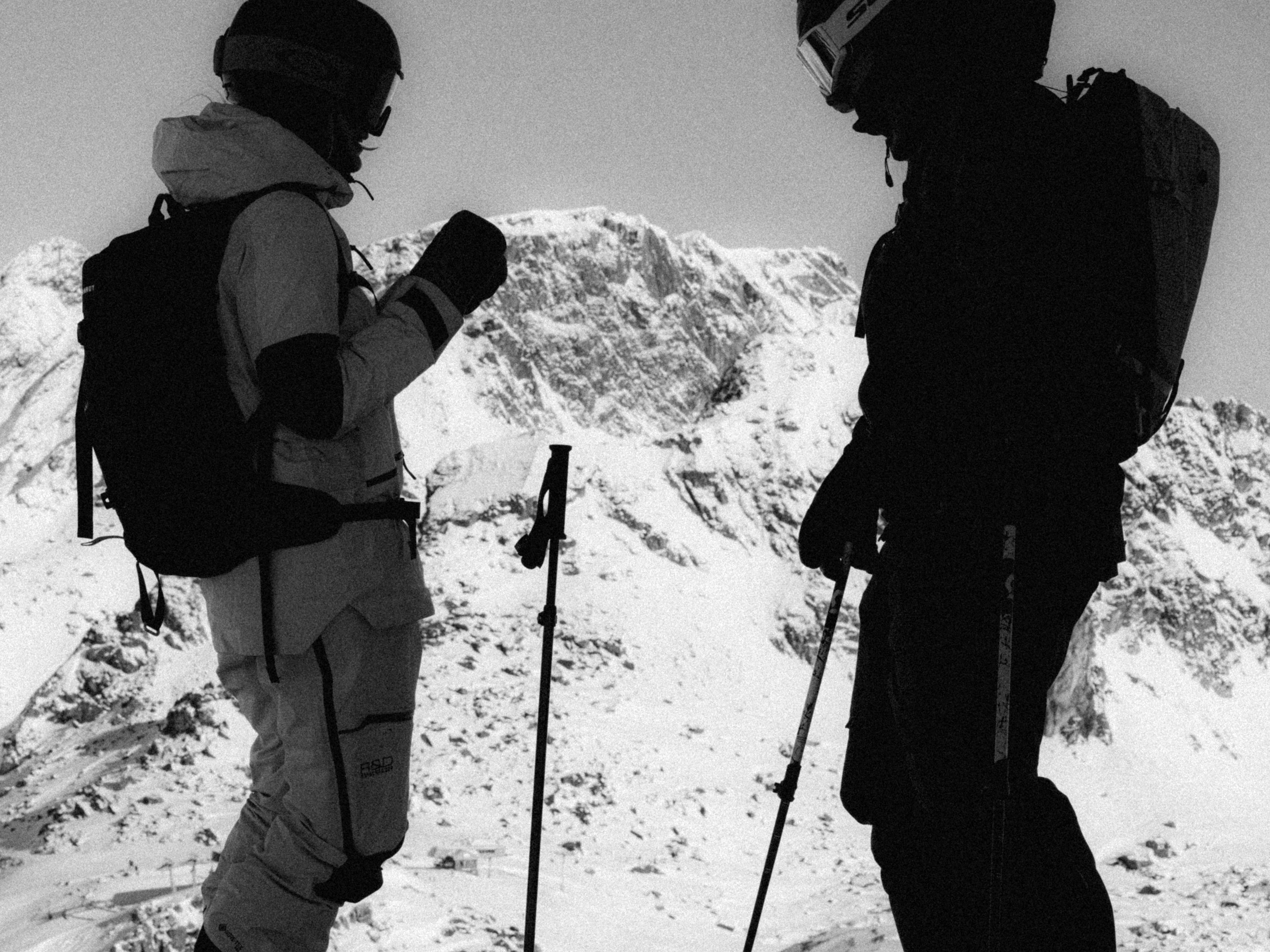 Black and white photo of two skiers standing on a snowy mountain ridge, one in light gray gear and one in dark silhouette, both carrying backpacks and holding ski poles.