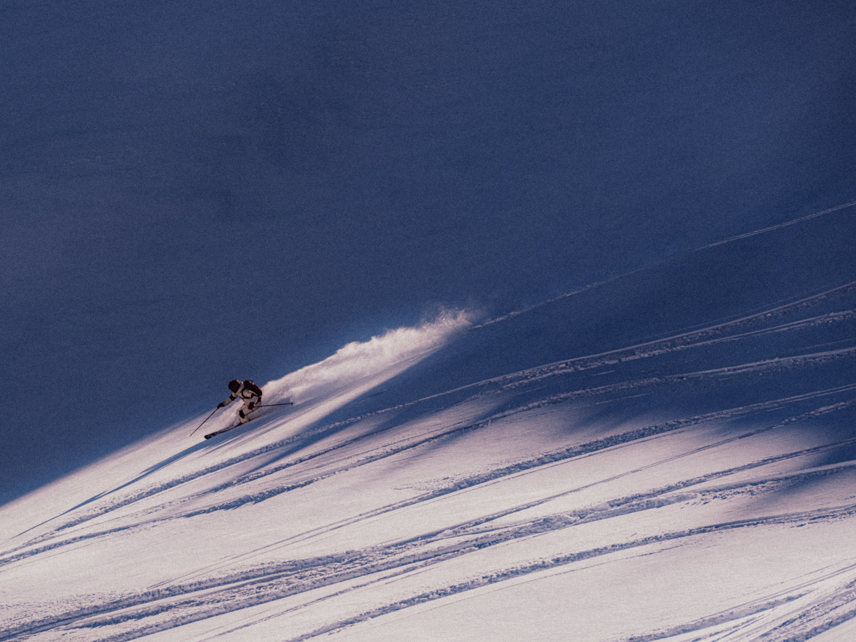 A skier carving through fresh powder snow on a steep slope under a deep blue sky, leaving a spray of snow behind them.
