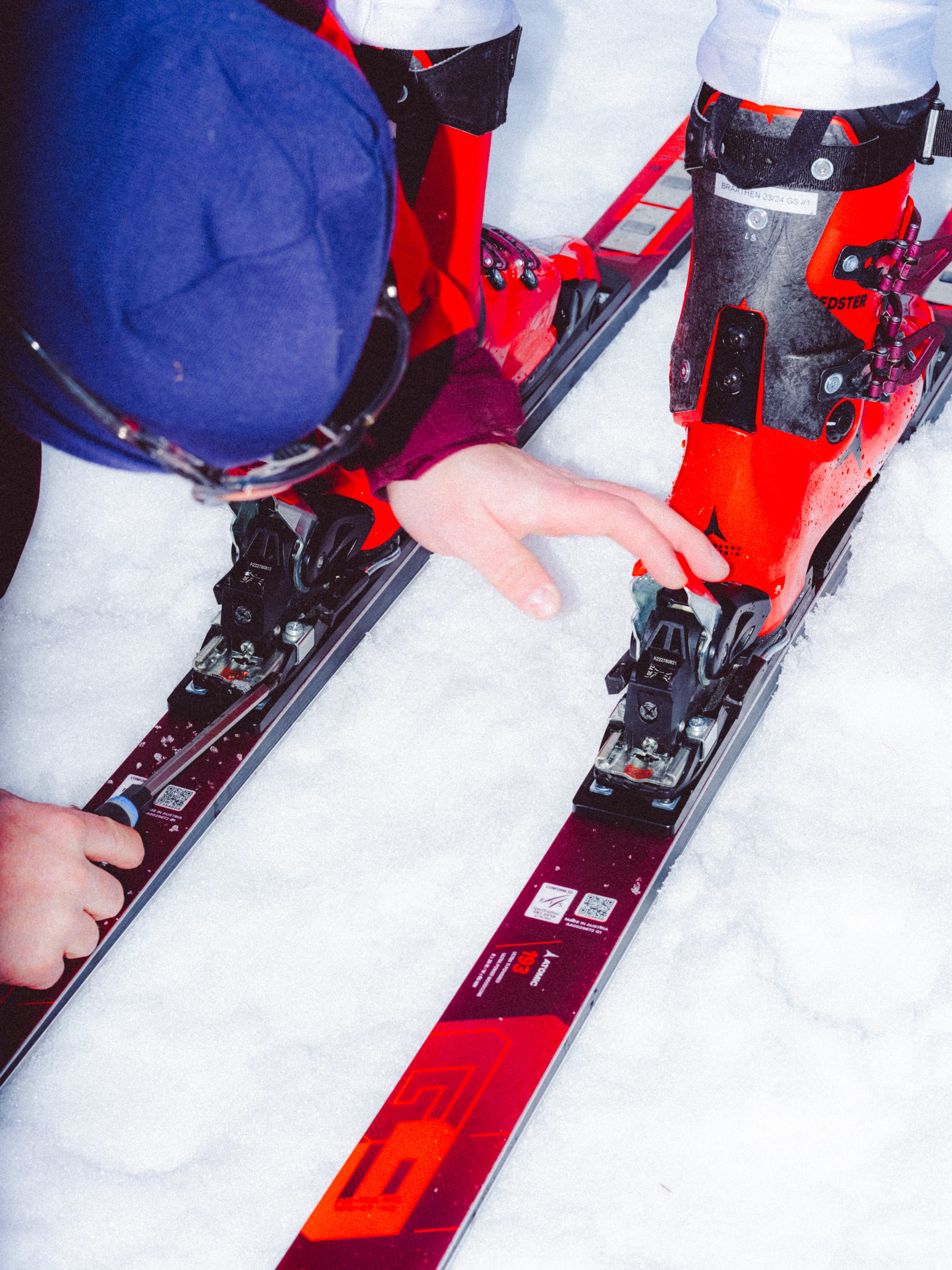 Close-up of a skier adjusting the bindings on red and black skis in the snow, wearing a blue helmet and red ski boots.