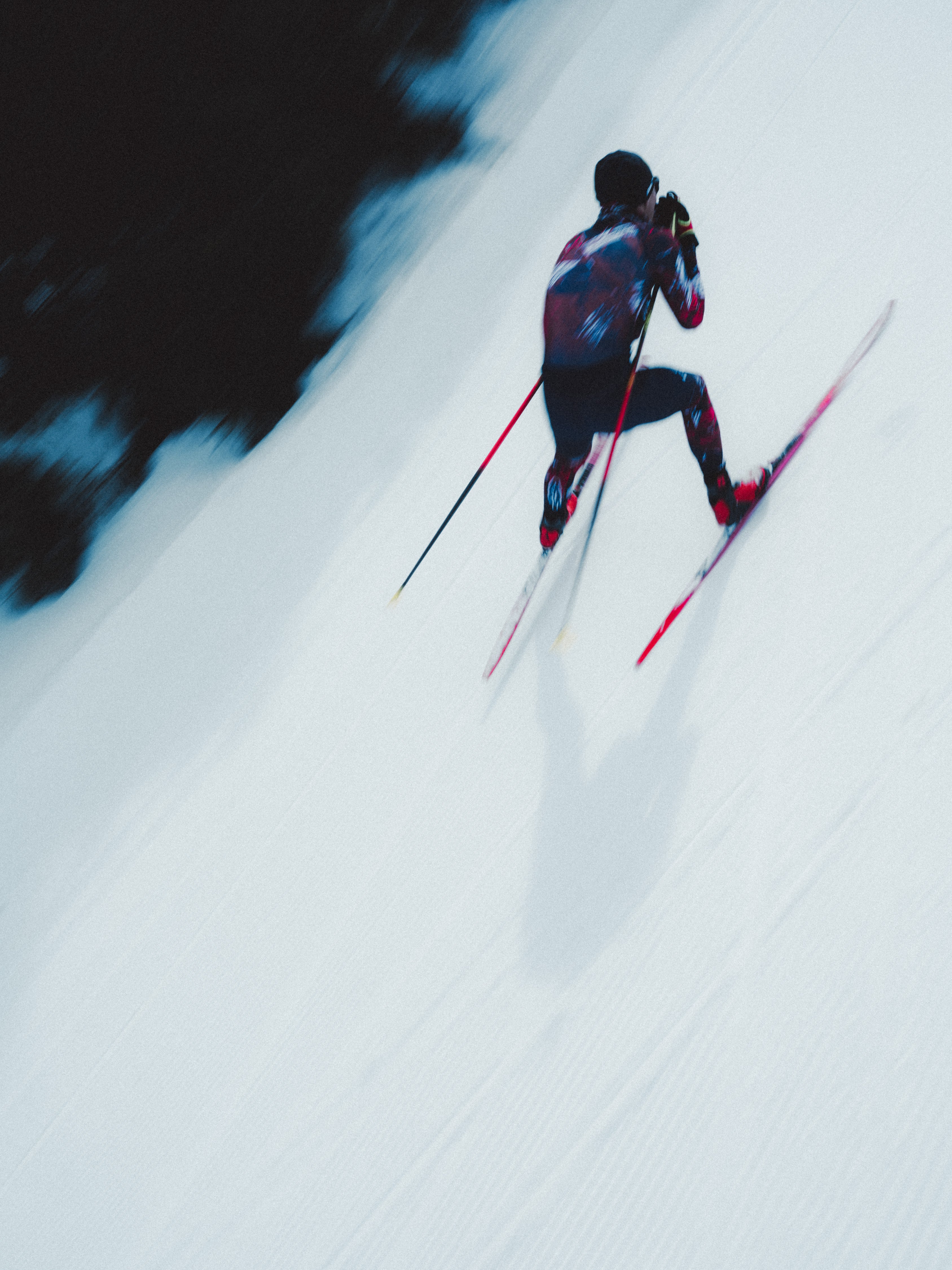 Mika Vermeulen in a dark racing suit with red and blue graphics glides down a snowy slope, holding ski poles and wearing red skis, captured from a high-angle perspective against a bright white snow background.