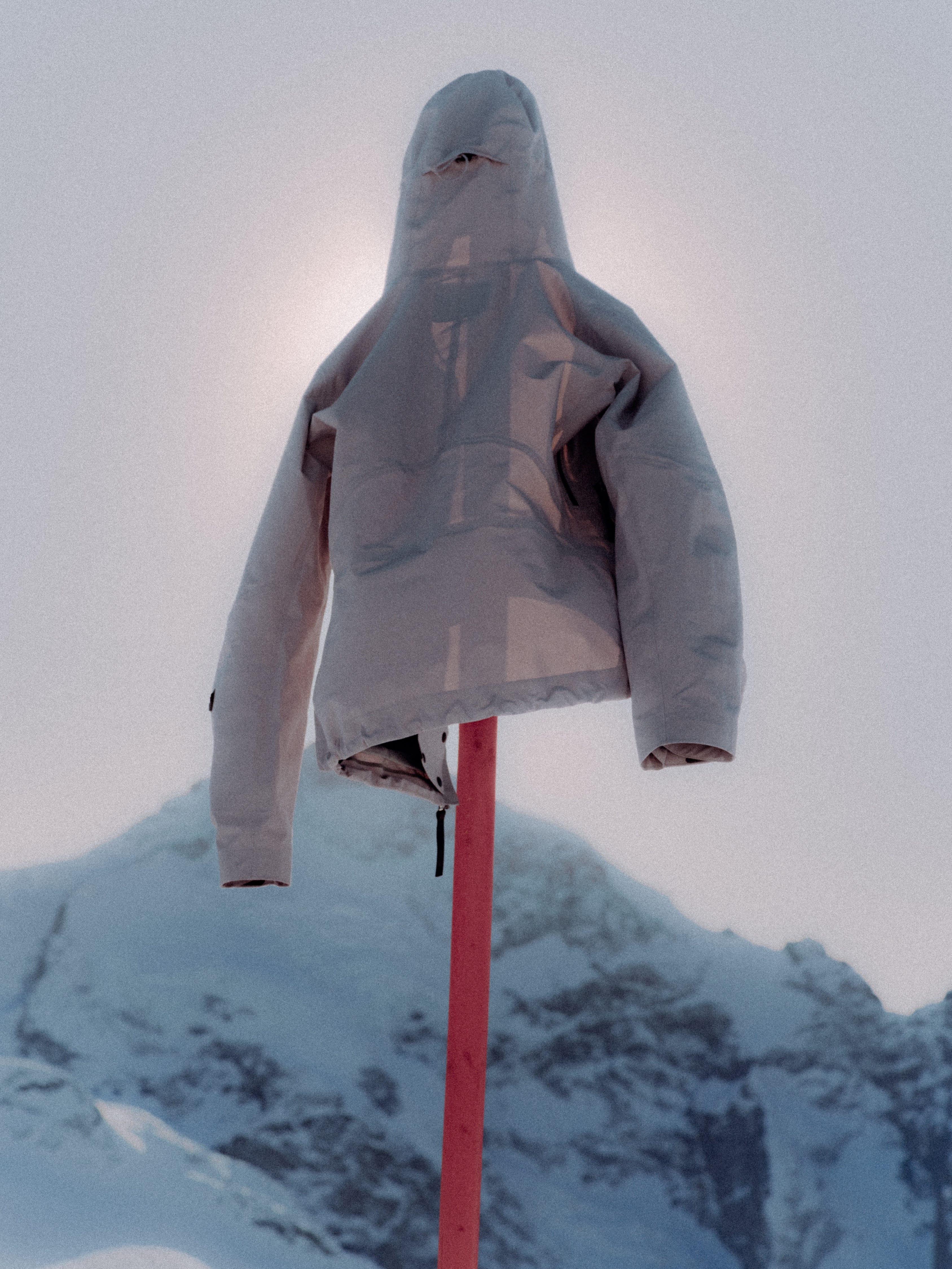 A light gray hooded jacket or cover draped over a red pole, photographed outdoors against a snowy mountain and pale sky.