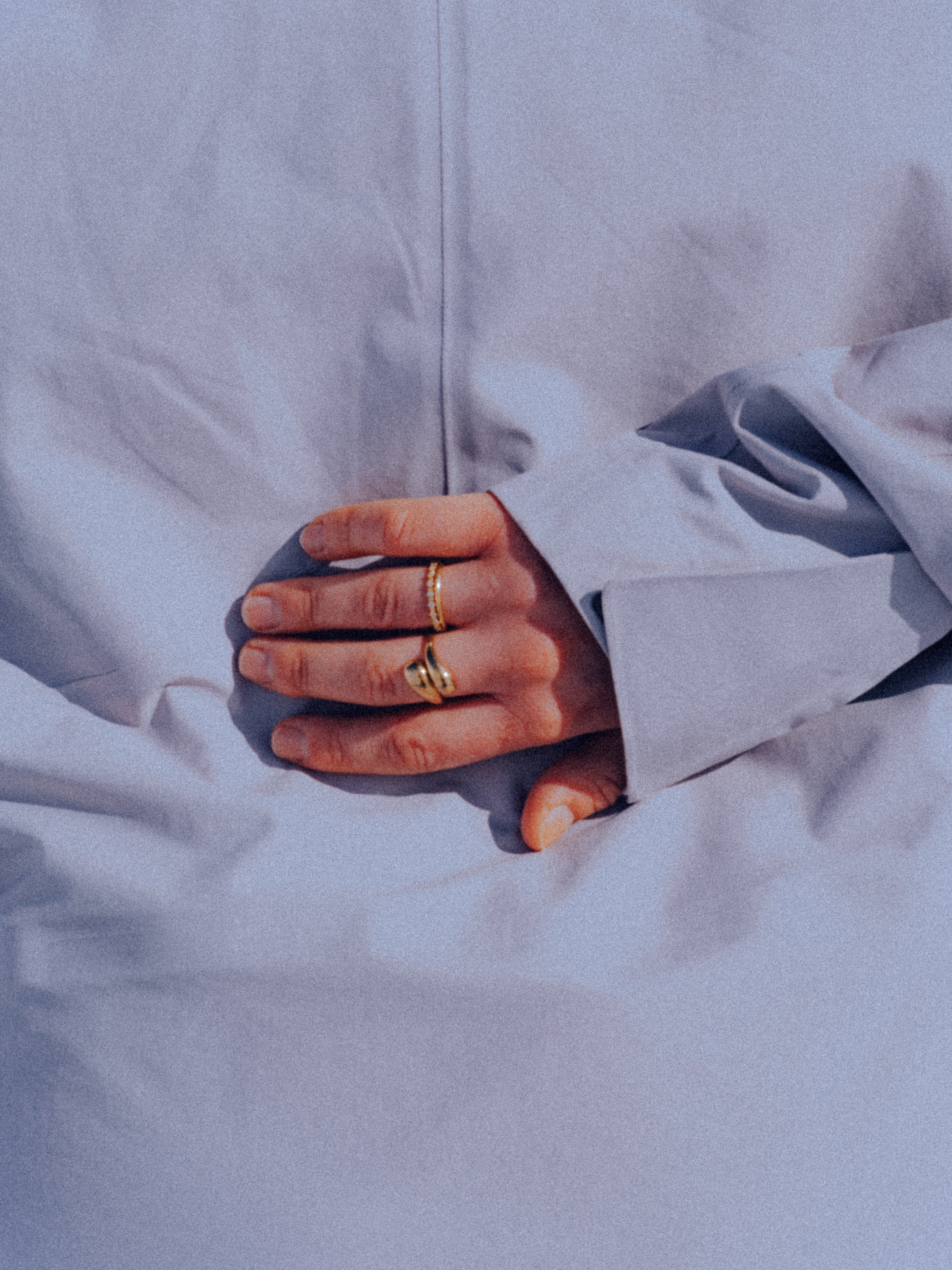 A close-up of a person's hand resting in soft light grey fabric, wearing two gold rings.