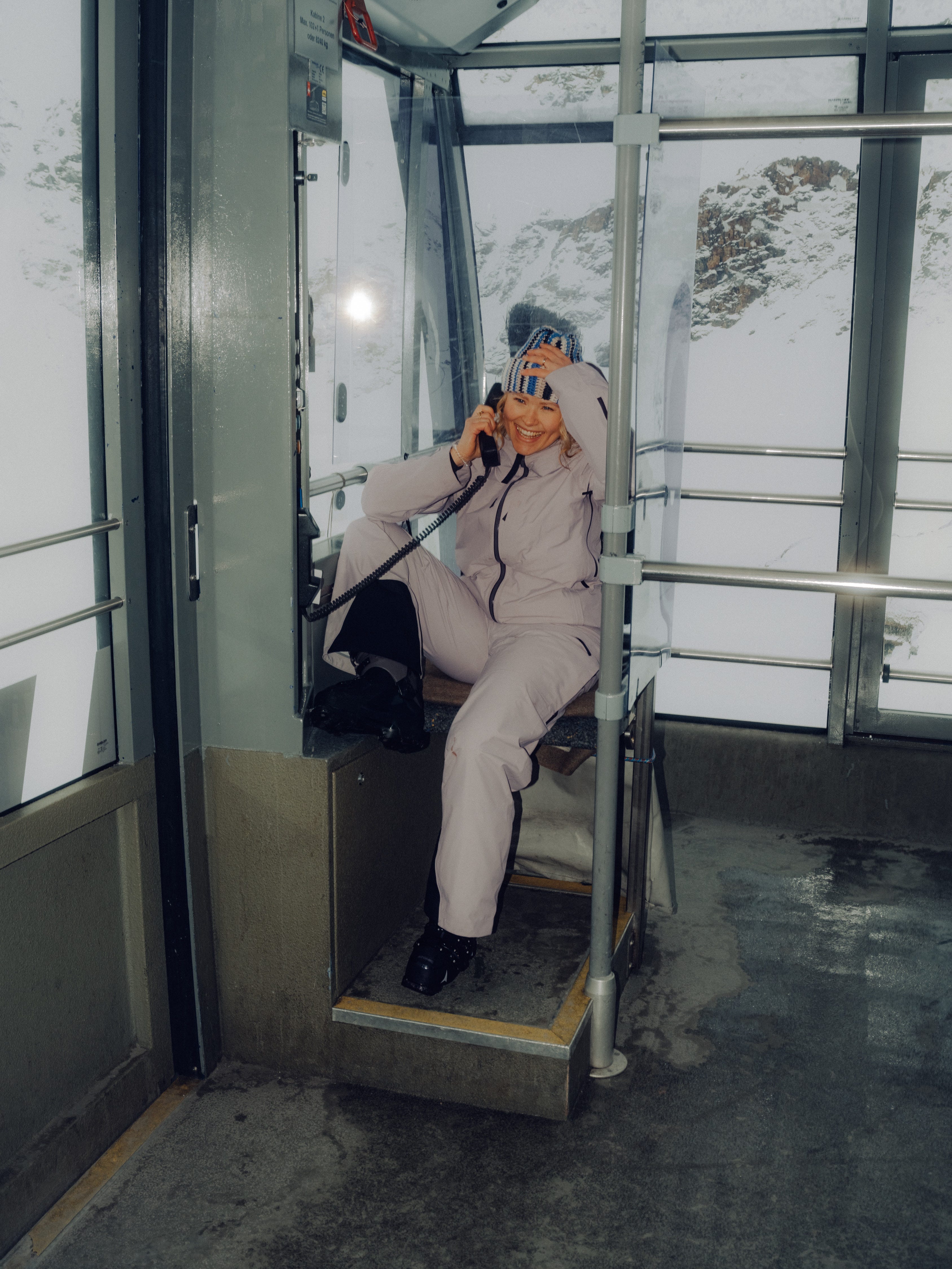 A person wearing a light grey ski suit and goggles sits inside a metal gondola lift cabin, holding a phone, with snowy mountains visible through the large windows.