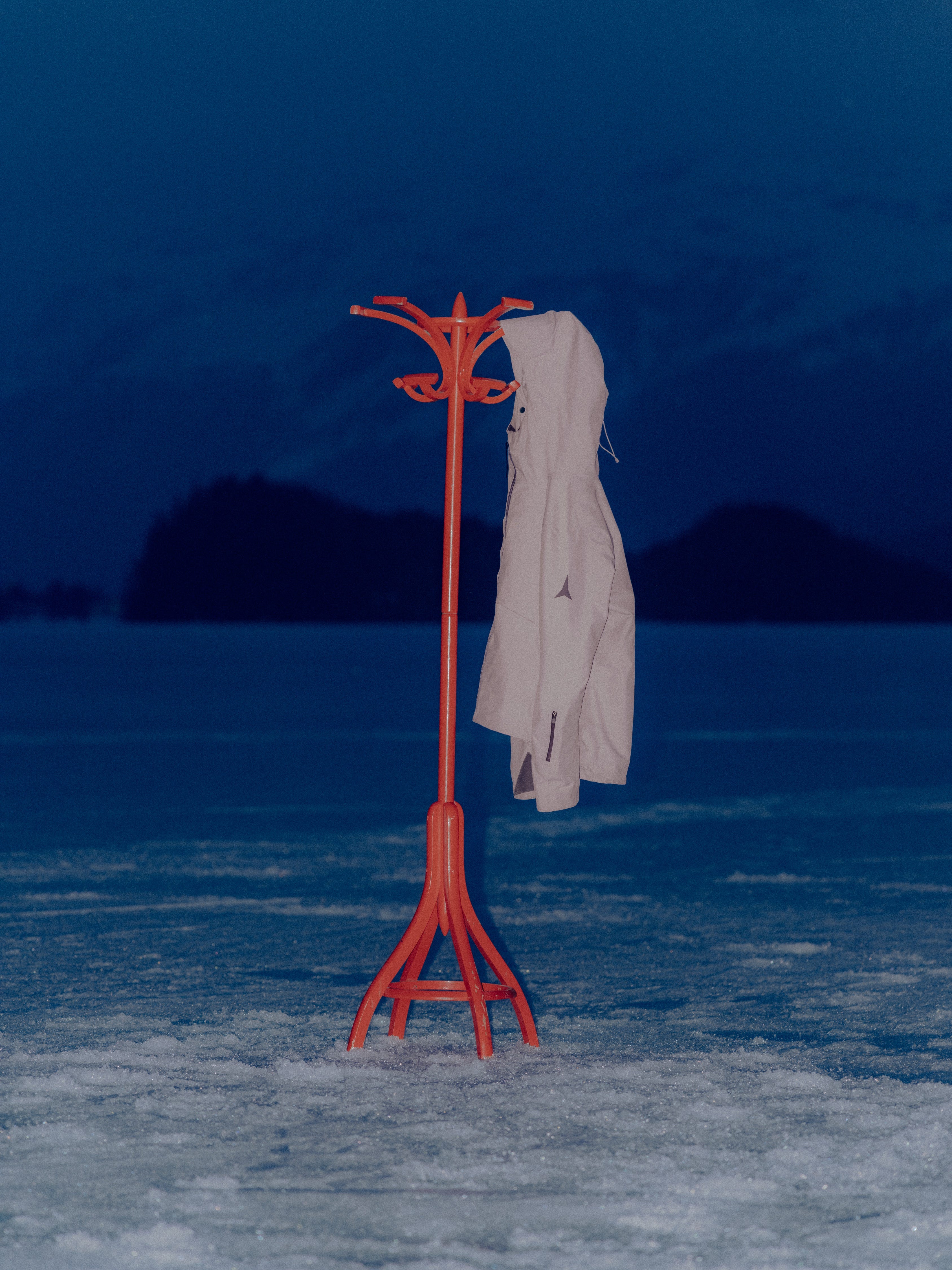 A red coat rack with a gray coat hanging on it on a snowy surface during the night. 