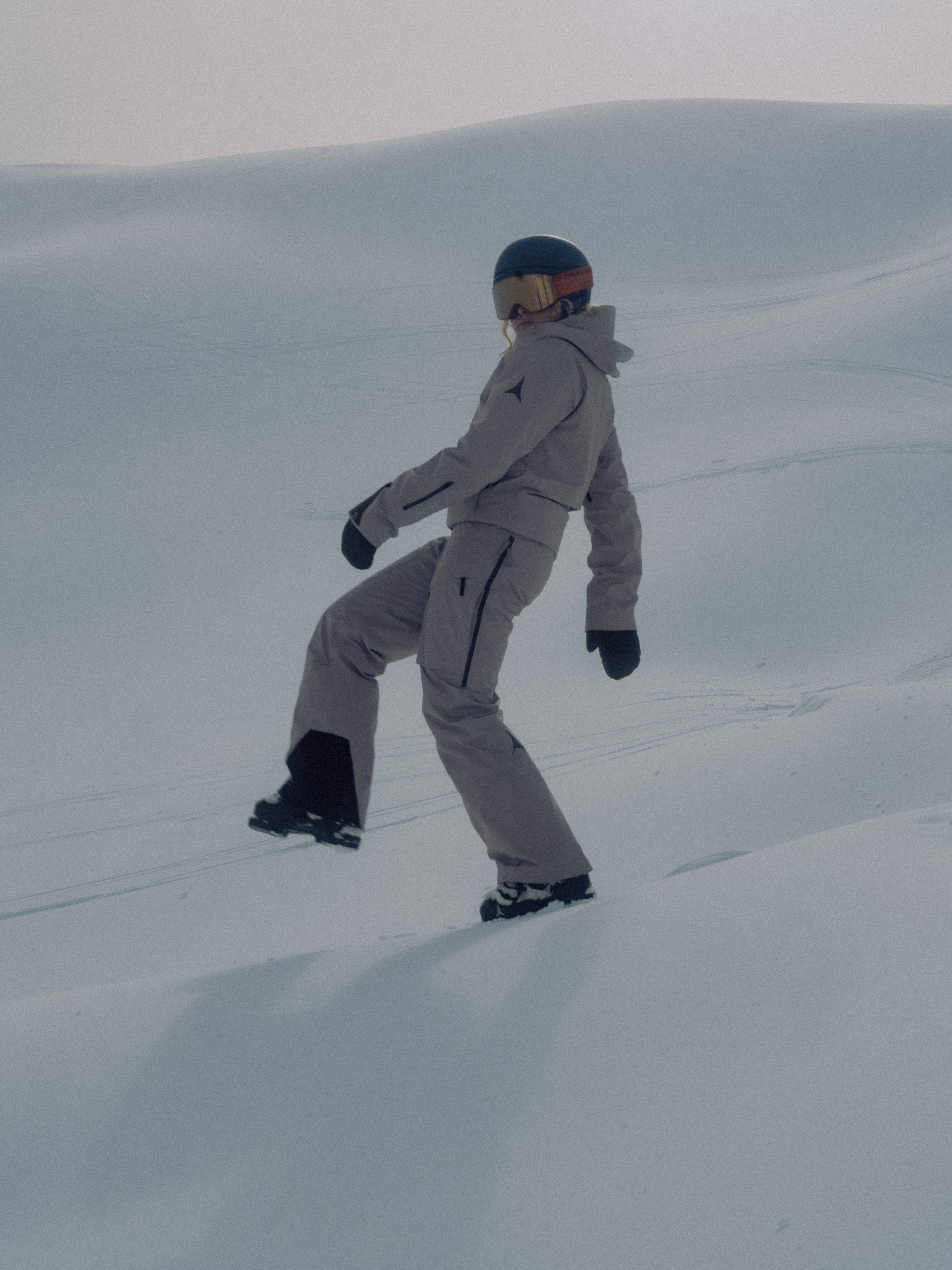 A person walking on a snowy slope, wearing a light gray ski suit, black helmet, and goggles.