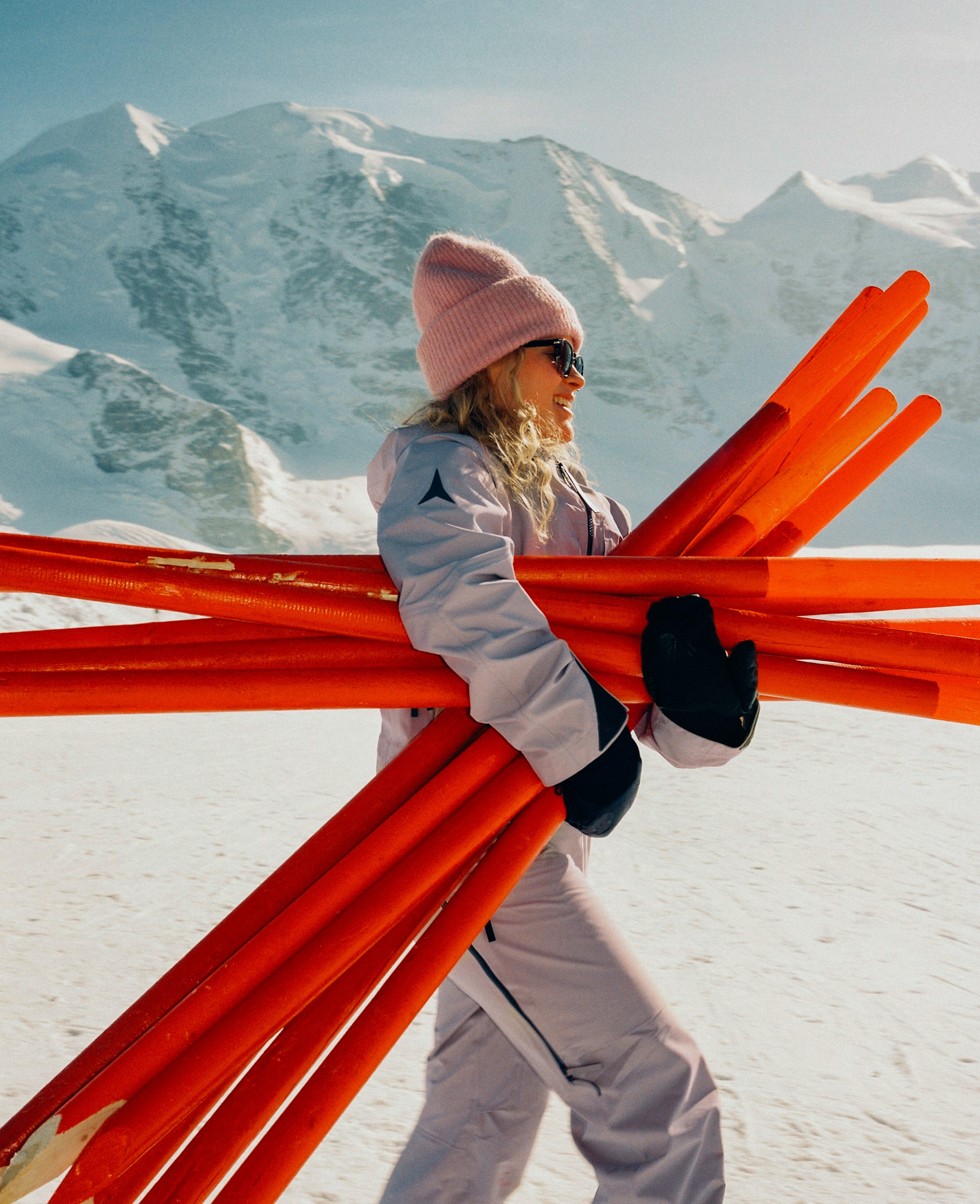 A woman in ski gear carrying a bundle of bright orange skis in a snowy mountain setting.