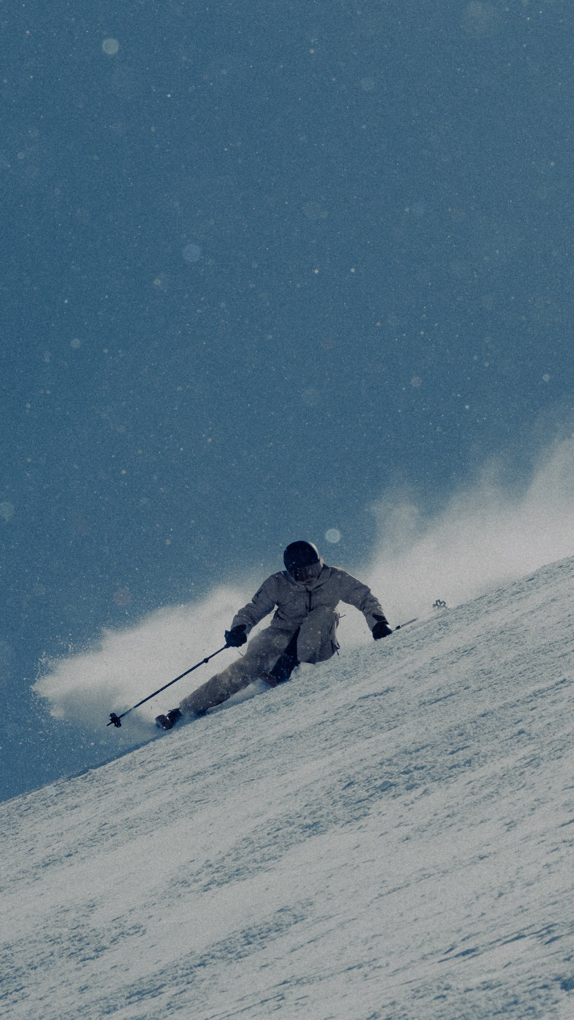 A skier in a light-colored snowsuit moves down a snowy slope on a blue, snowy day, kicking up a spray of snow.
