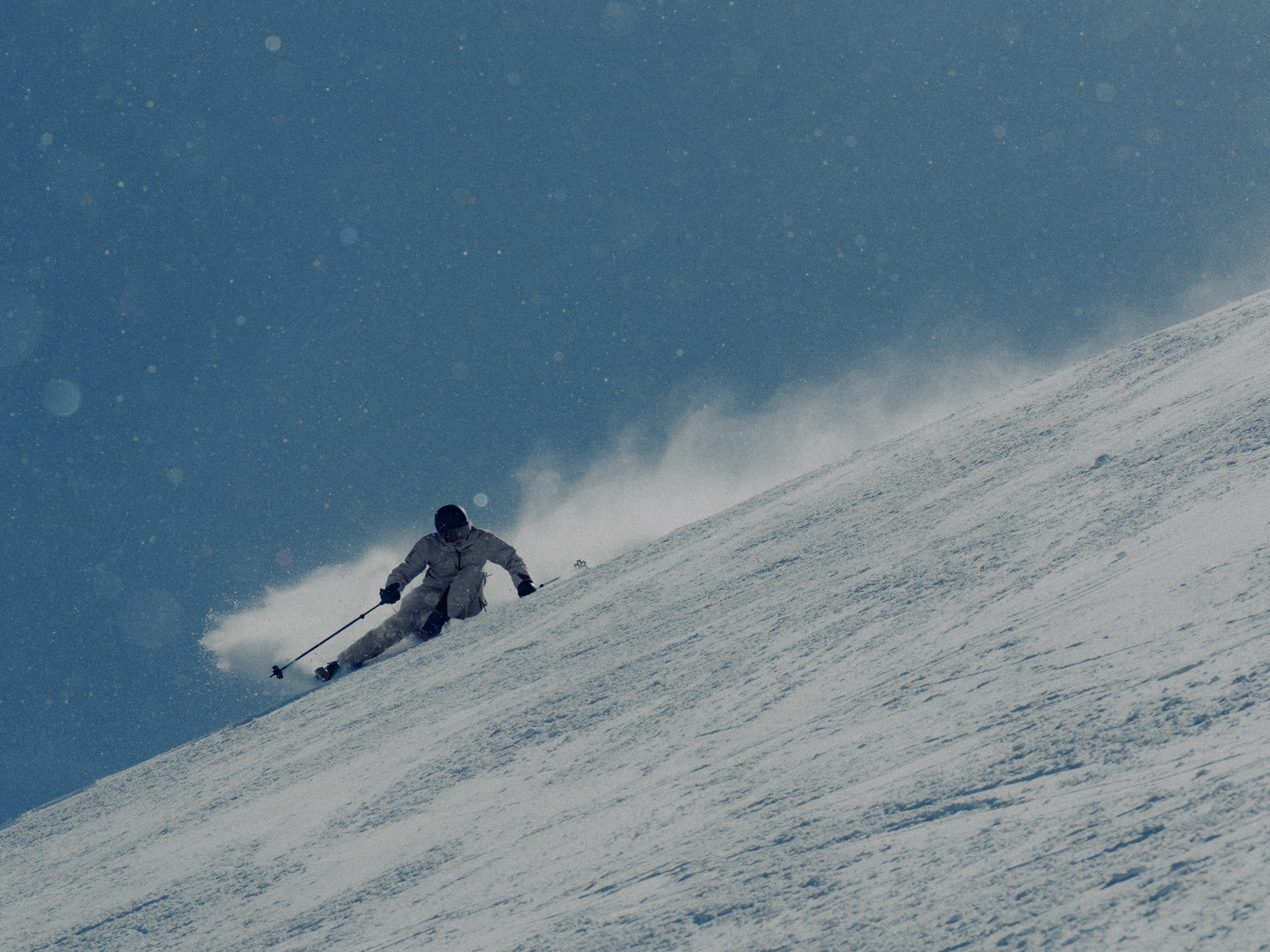 A skier in a light-colored suit carving through fresh powder on a snowy slope, with a spray of snow kicked up behind them under a blue sky.