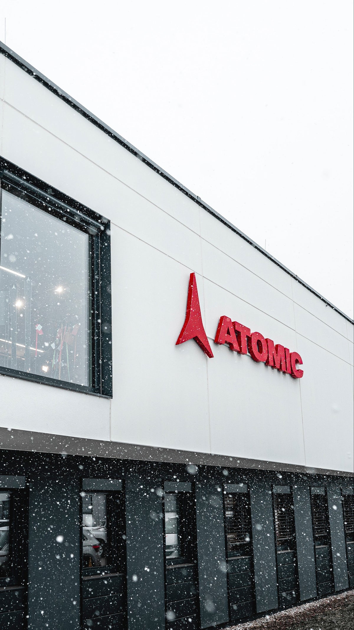 A modern white and black building exterior with a prominent red ATOMIC logo on the wall, photographed in falling snow under an overcast sky.