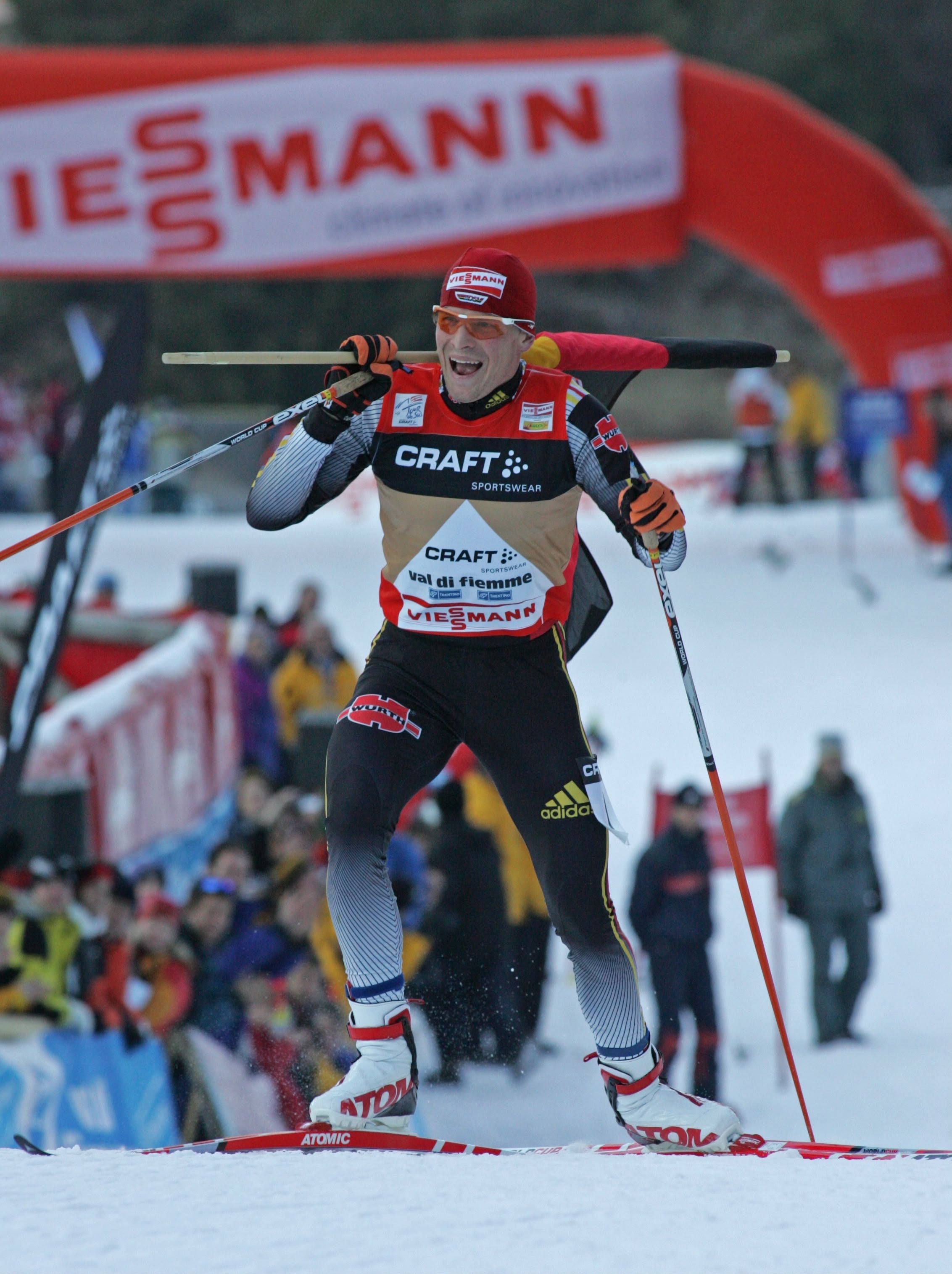 Tobias Angerer in a black, red, and beige racing suit with 'CRAFT' and 'adidas' branding, skiing through the snow during a race with a red archway and spectators in the background.