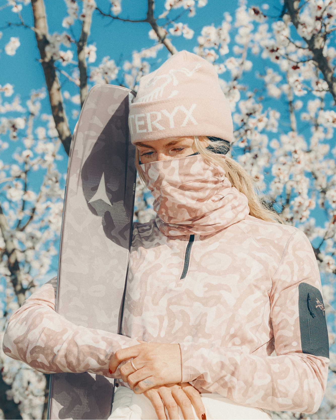 Tonje Kvikik wearing a light pink patterned ski jacket, a matching beanie with 'EVERYX' printed on it, and a face covering, holding a dark ski board against a background of blooming white trees and blue sky.