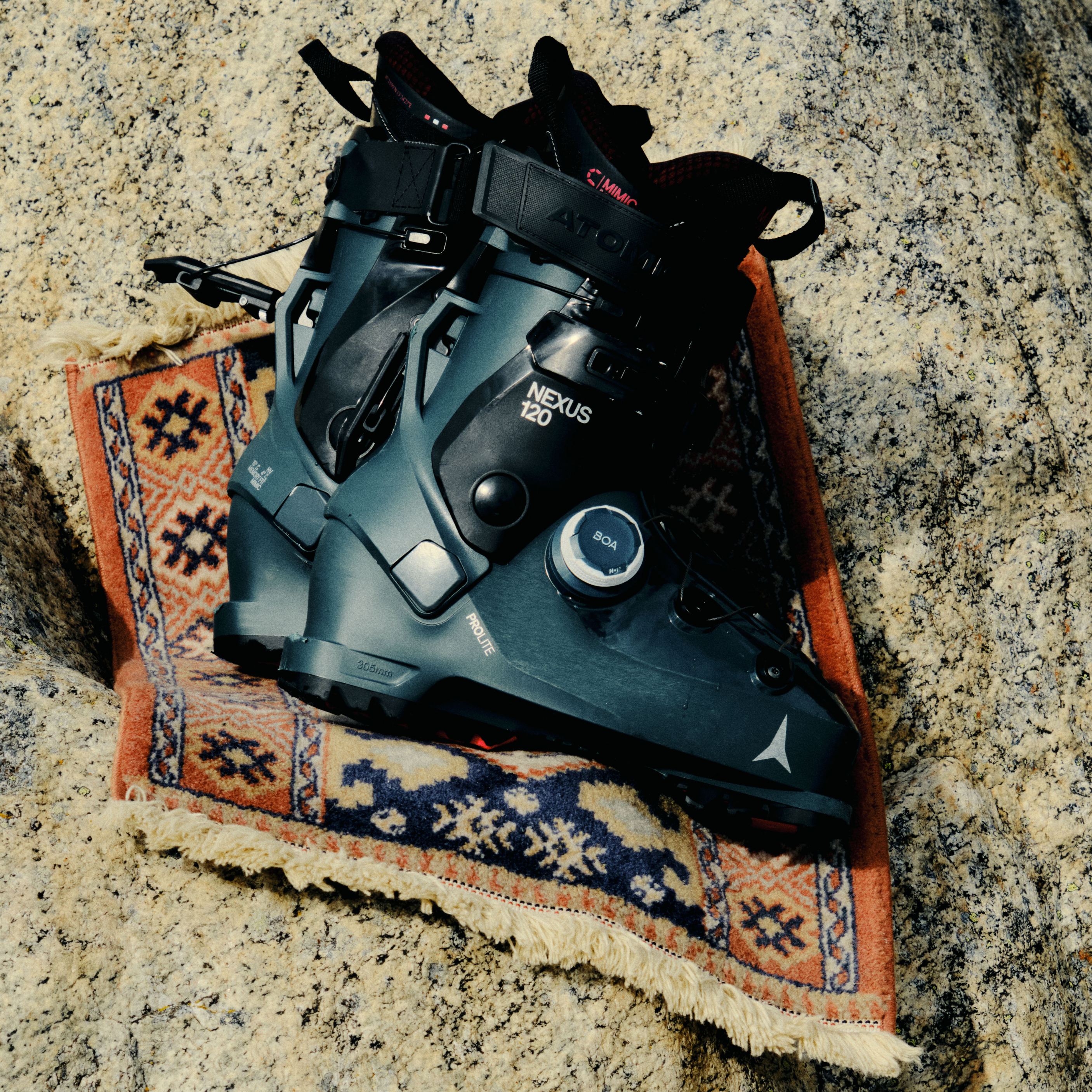 A pair of black and dark gray ski boots with white 'NEXUS 60' branding, resting on a small patterned rug over a textured stone surface.