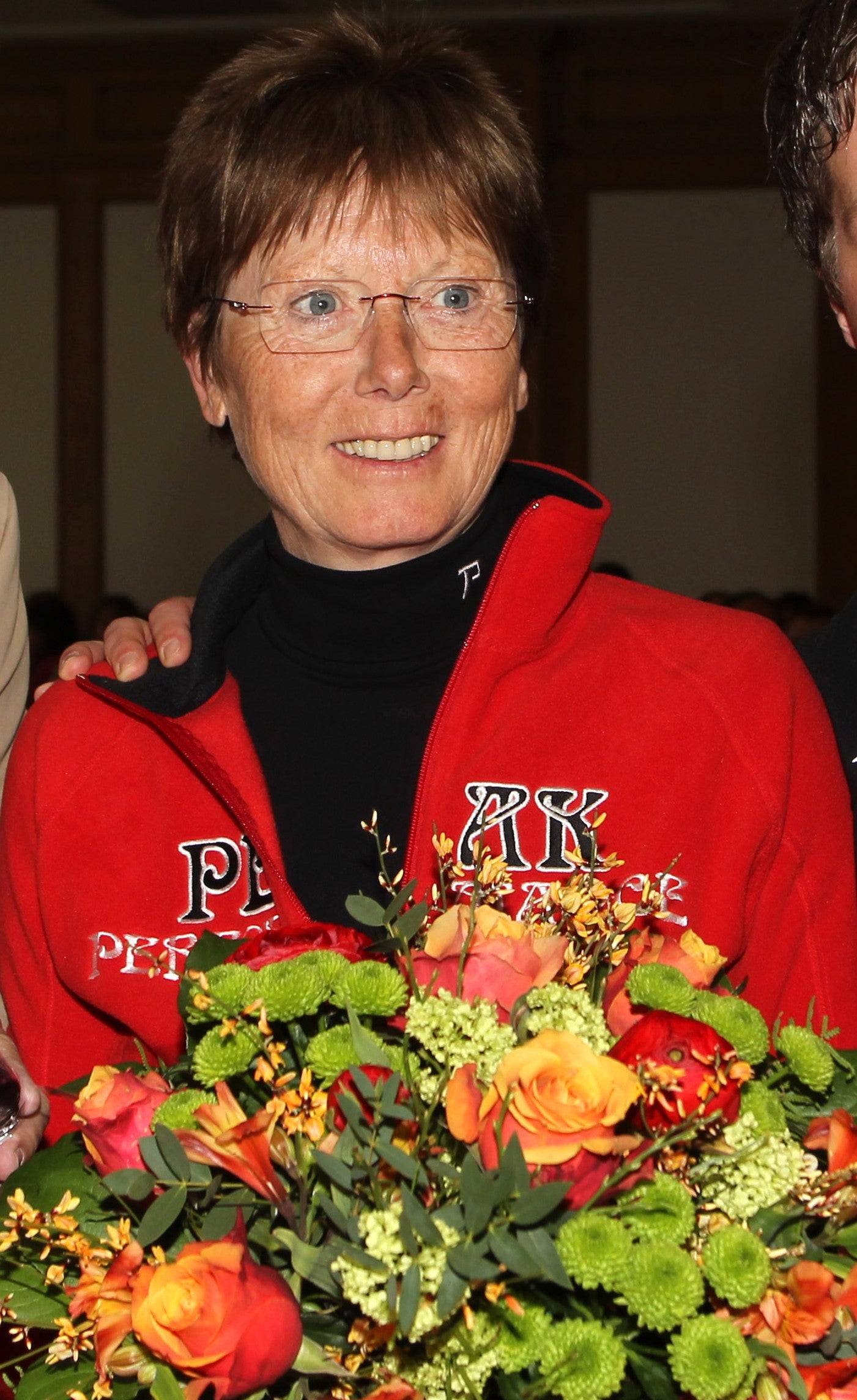 Annemarie Moser Proell with short brown hair and glasses, wearing a red zip-up jacket with 'PK' and 'PFF' embroidery over a black turtleneck, holding a large orange and green floral bouquet.