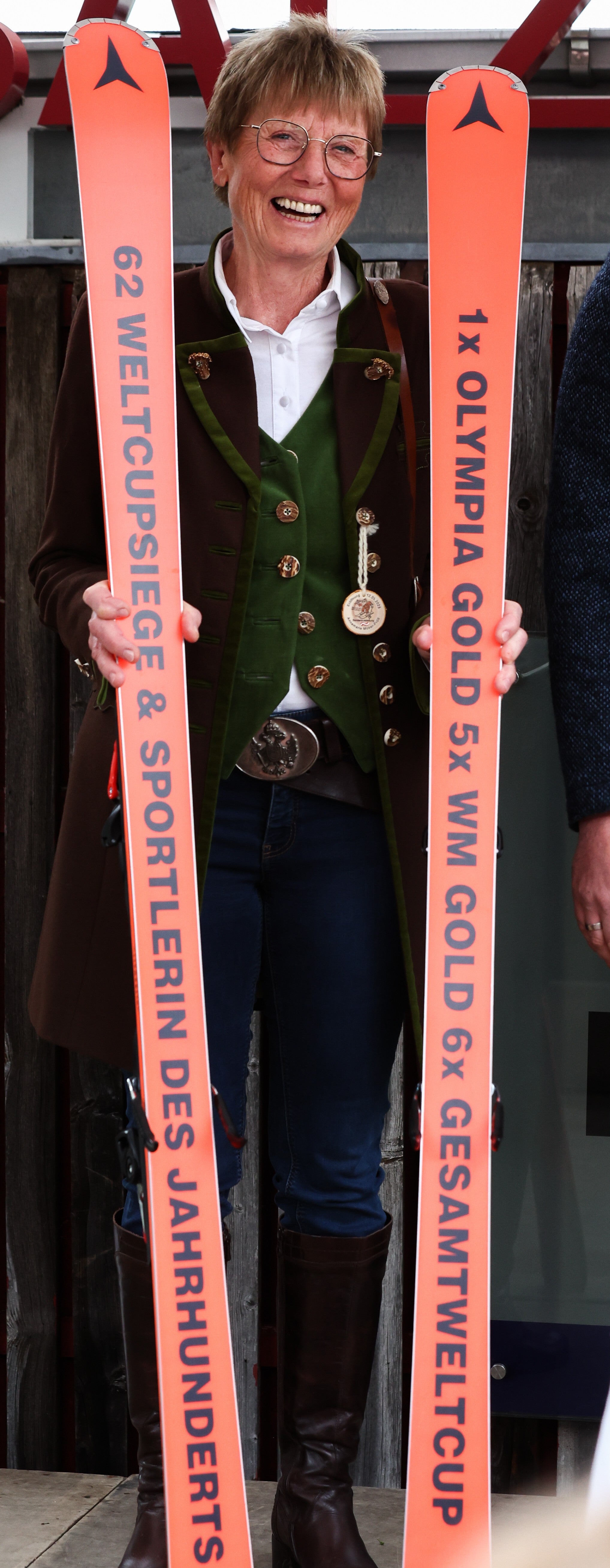 Annemarie Moser Proell in traditional Austrian clothing holds two orange skis printed with text celebrating the 62nd World Cup and Sportlerin des Jahrhunderts and the 1st Olympia Gold 5x WM Gold 6x Gesamtweltcup.