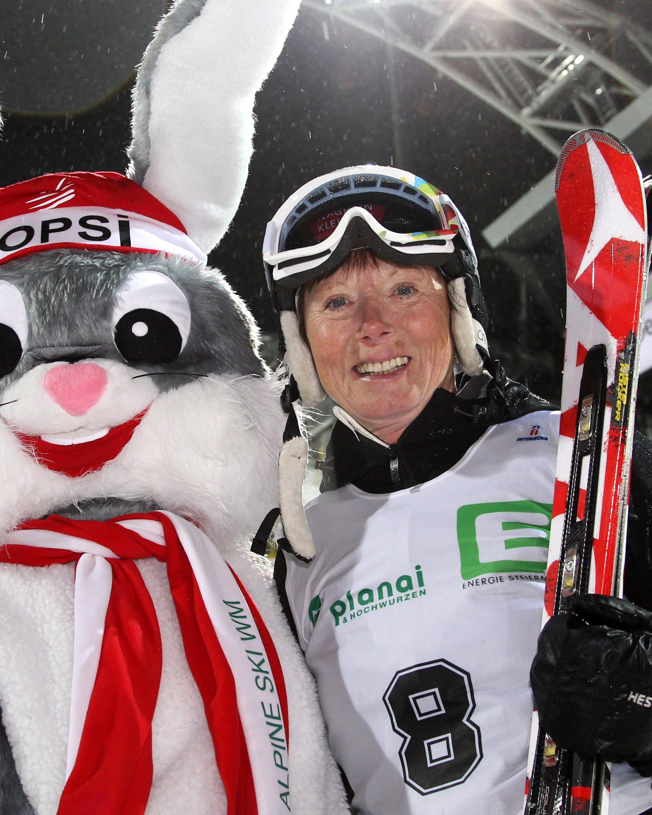 Annemarie Moser Proell in a white number 8 race jersey stands next to a large gray and white bunny mascot wearing a red and white 'Hopsi' headband and scarf, holding red and white skis at night under stadium lights.