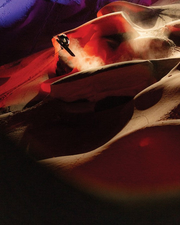 Chris Benchetler descending a dramatic night slope dramatically lit in deep reds and oranges, kicking up a cloud of powder snow against sculpted snow formations that resemble a slot canyon.