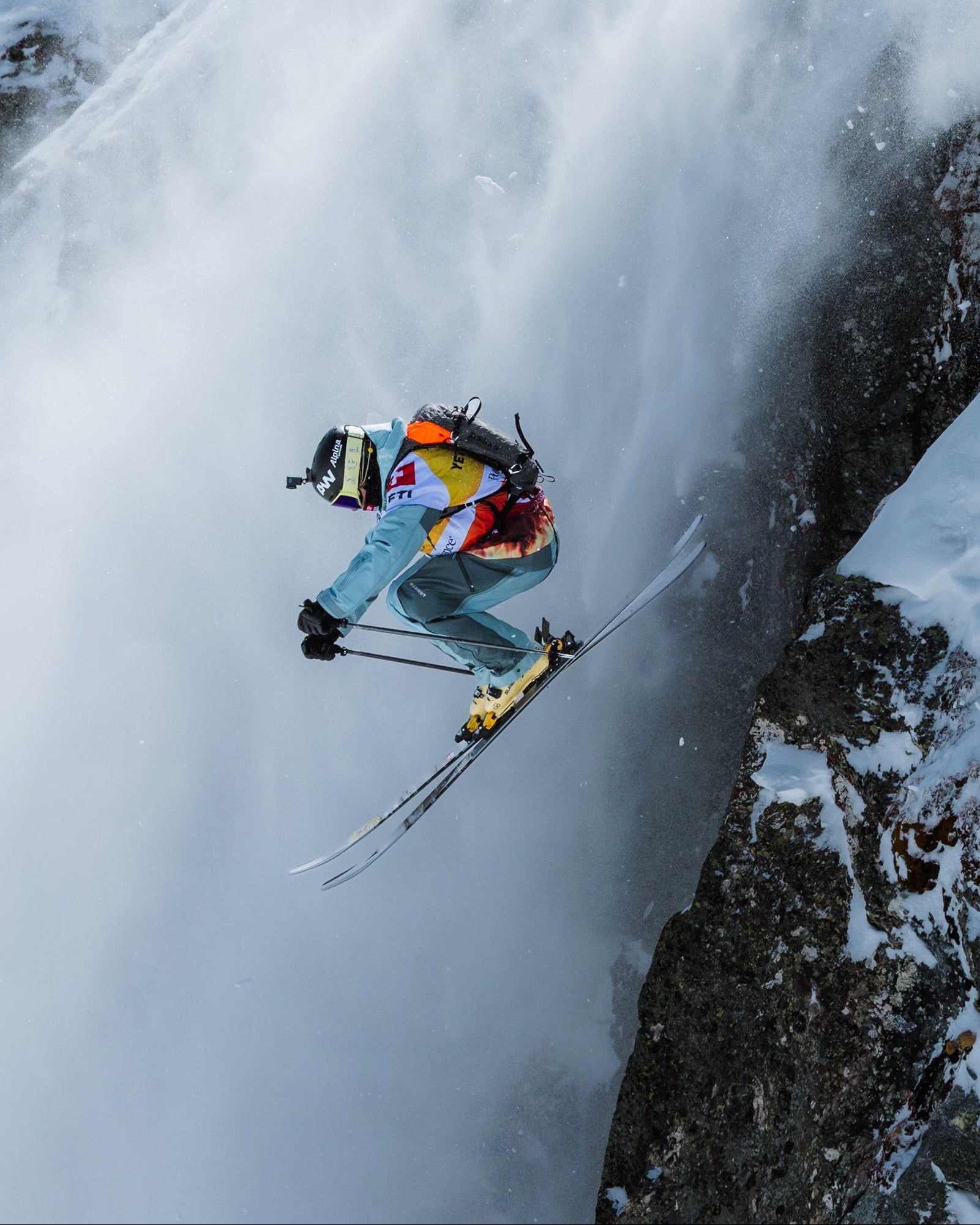 Sybille Blanjean in a colorful ski suit and helmet jumps through the air beside a steep snowy mountain wall with exposed rock and a large plume of snow spray.