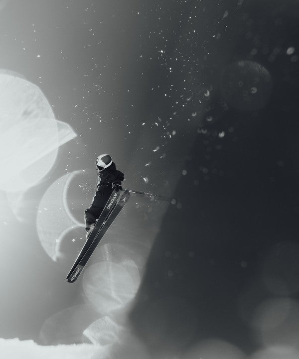 Fabian Boesch in dark winter gear is captured mid-air against a dramatic black-and-white snowy background with soft bokeh light circles.