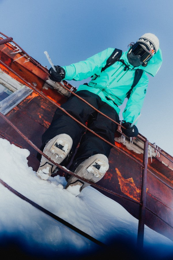 Bobby Brown in a bright turquoise ski jacket, black ski pants, and white ski boots sits on a rusty red metal structure in the snow under a clear blue sky.