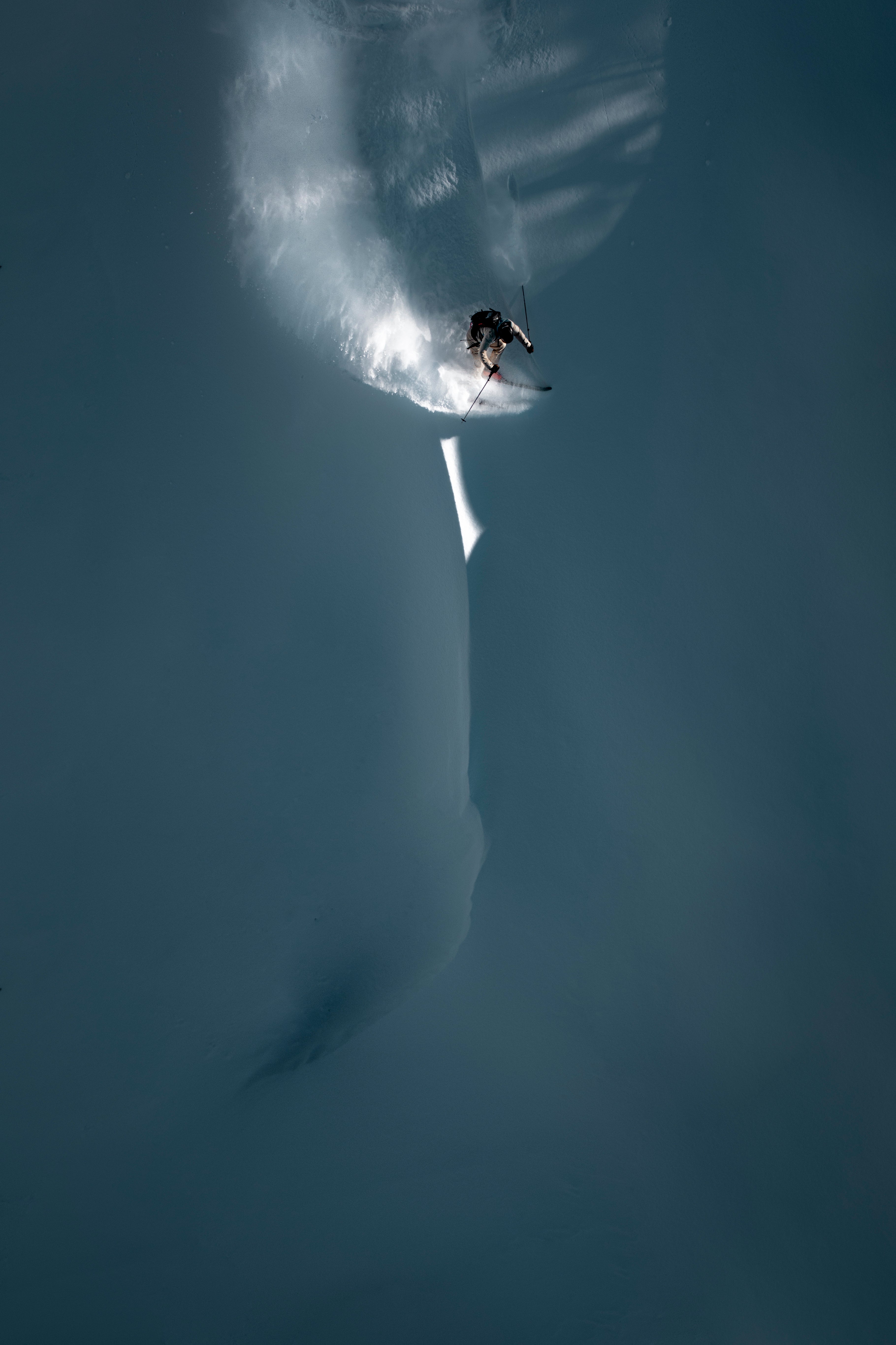 Matthias Haunholder carving through deep powder snow, creating a bright spray of powder and leaving a long shadow behind them in a dark blue-gray landscape.