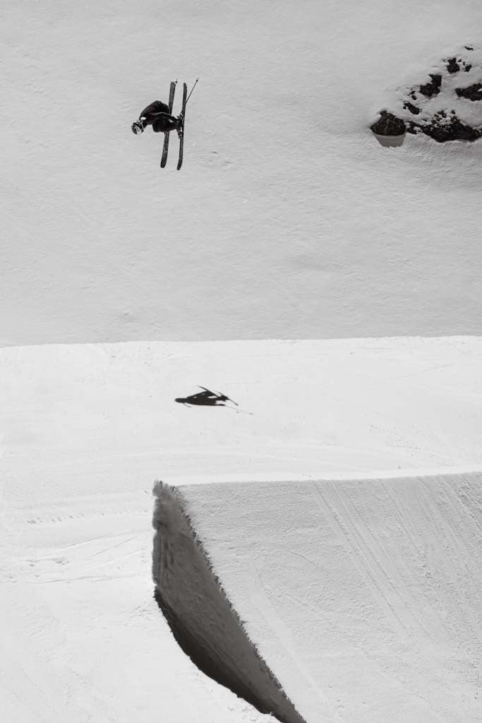 A black-and-white action shot of Tess Ledeux mid-air performing a trick above a snowy halfpipe, with another skier lying on the snow below.