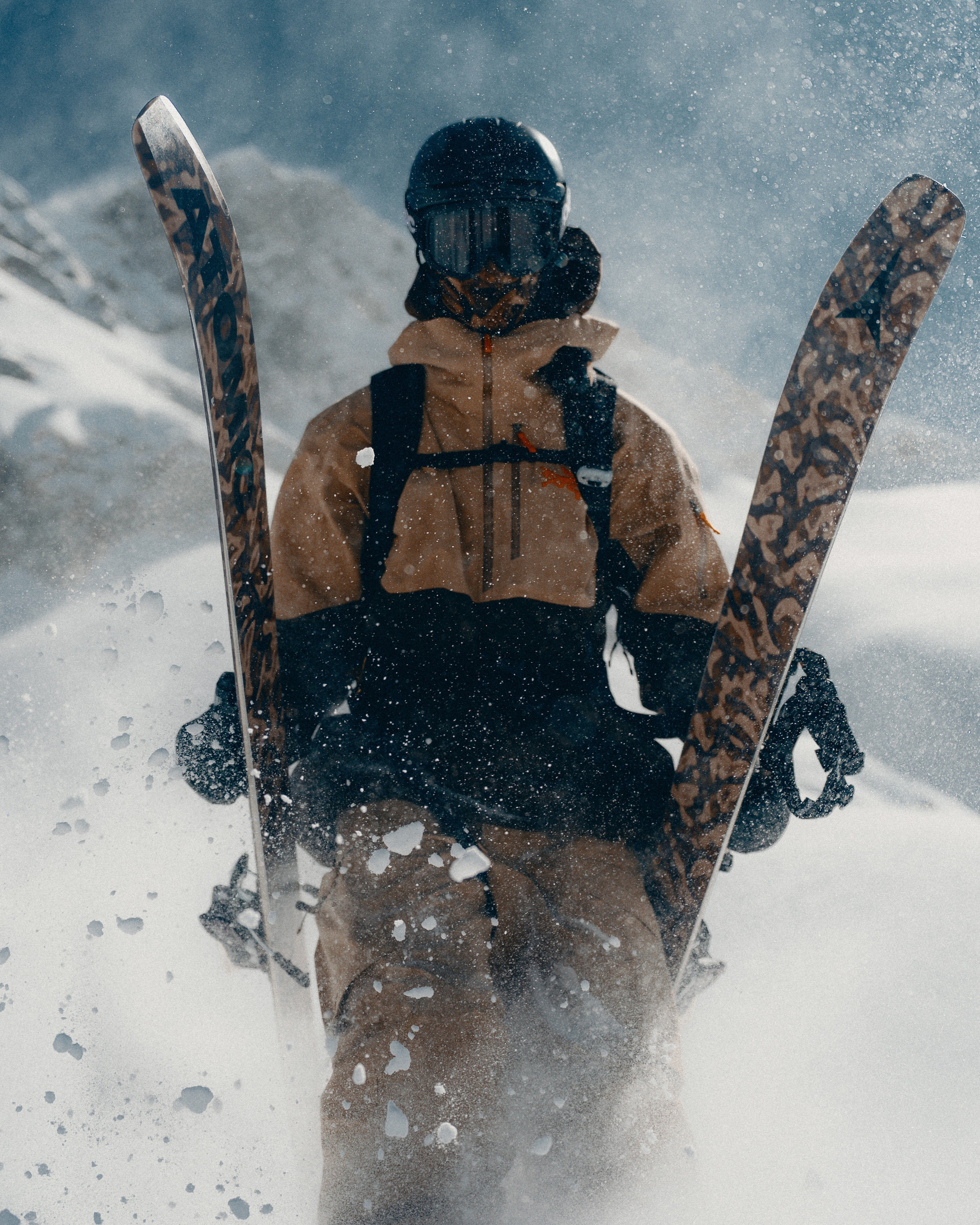 Craig Murray in a ski outfit stands in deep snow with skis raised behind them. The outfit is tan and black, and the scene is covered in snow with visible spray and a hazy outdoor background.