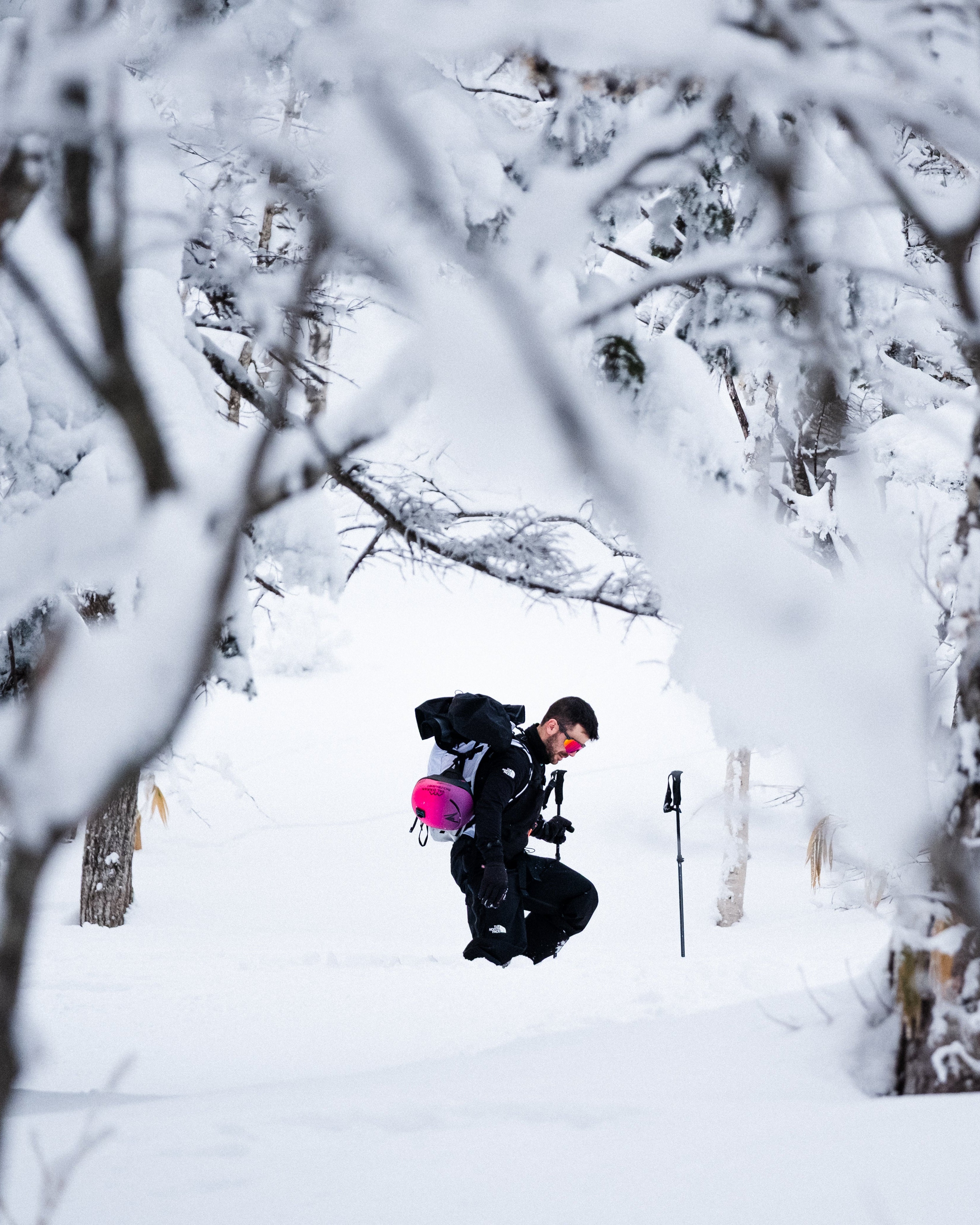 Aymar Navarro in black winter clothing with a bright pink backpack stands in deep snow among snow-covered trees, holding a trekking pole.