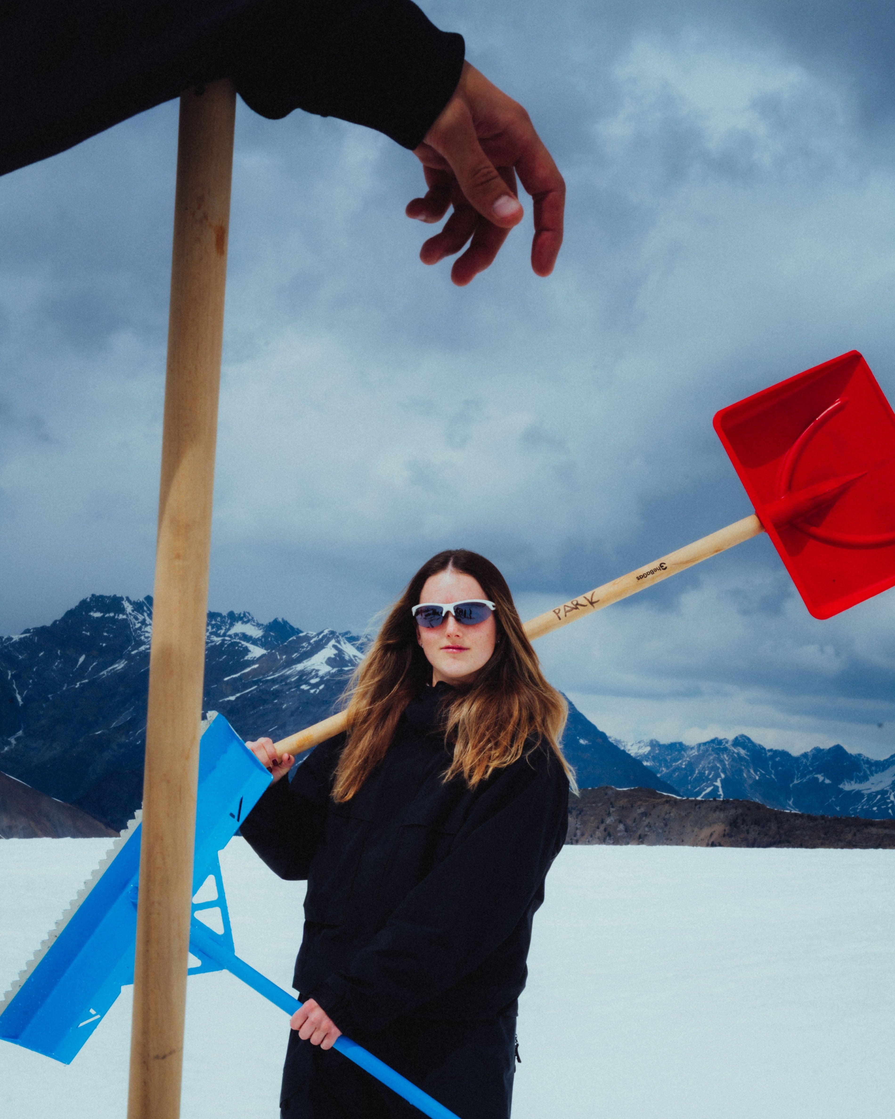 Megan Oldham in a black winter outfit and sunglasses stands on a snowy landscape with mountains in the background, holding a blue snow shovel and a red snow shovel over her shoulder. A large wooden pole and a hand at the top frame the shot.