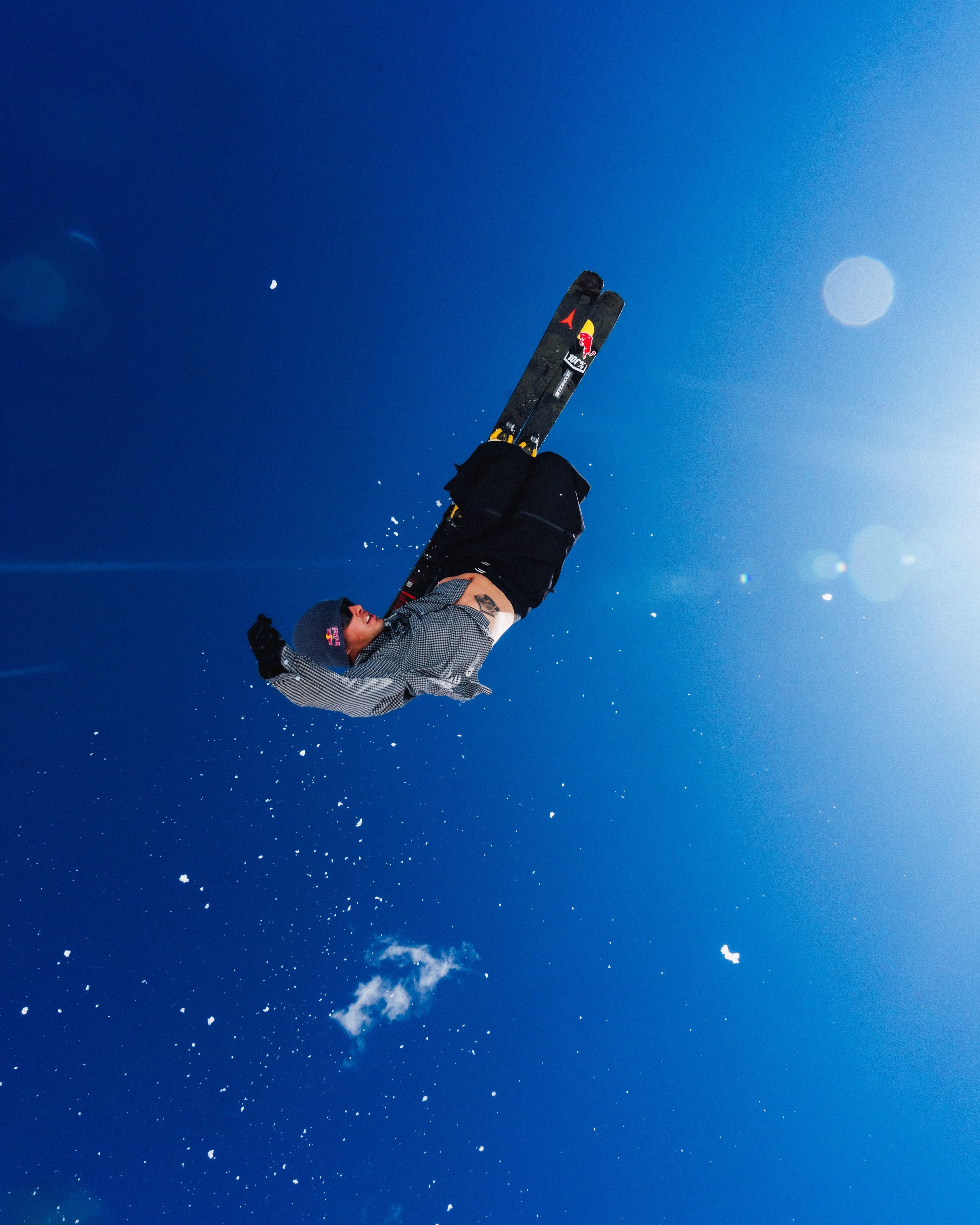 Nico Porteous is captured mid-air performing a high jump or trick against a bright blue sky with sun flares. The skier is wearing a gray jacket, black pants, and black skis with red and yellow bindings.