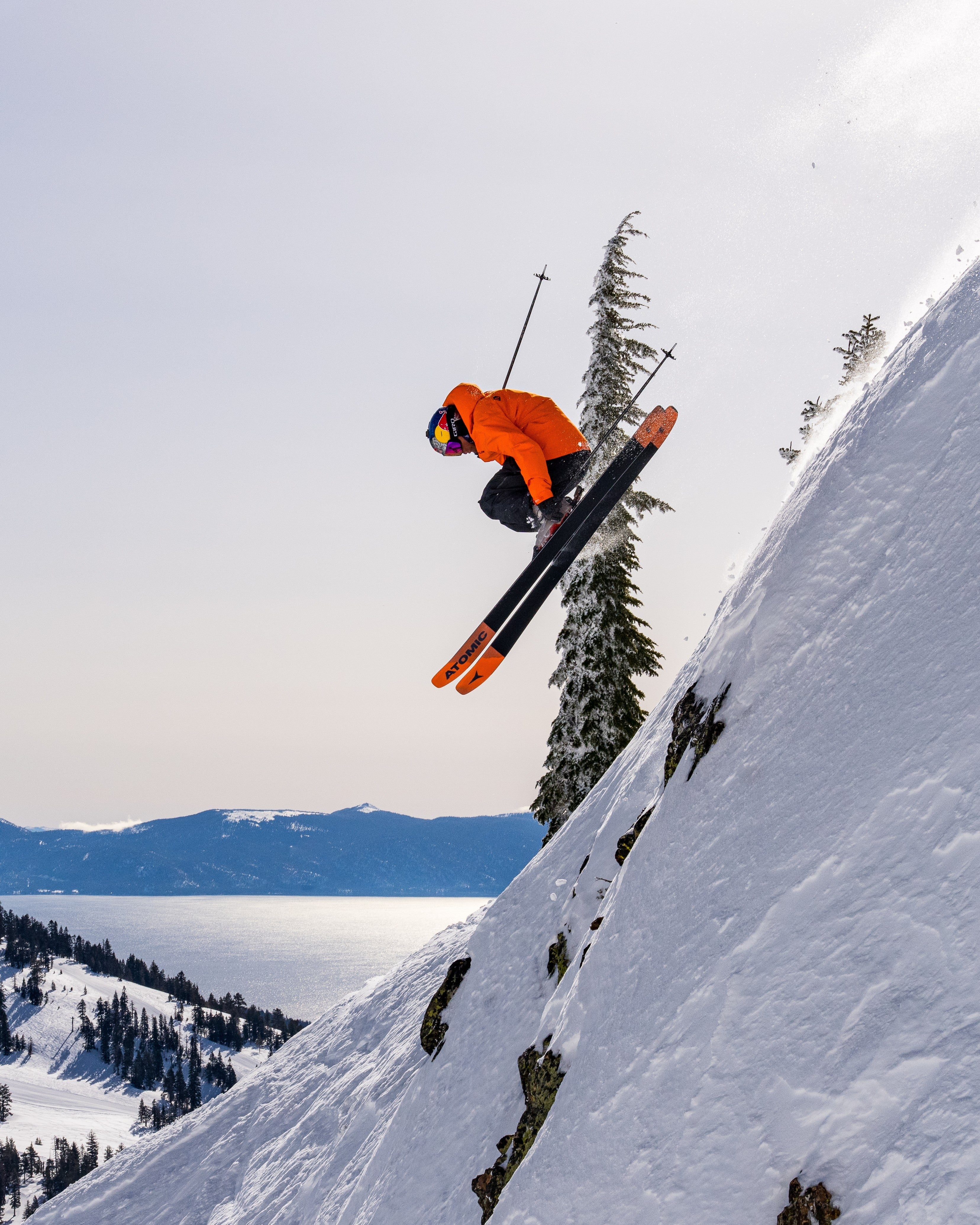 Daron Rahlves in an orange jacket mid-jump on a snowy mountain slope, with trees, distant mountains, and a pale sky in the background.