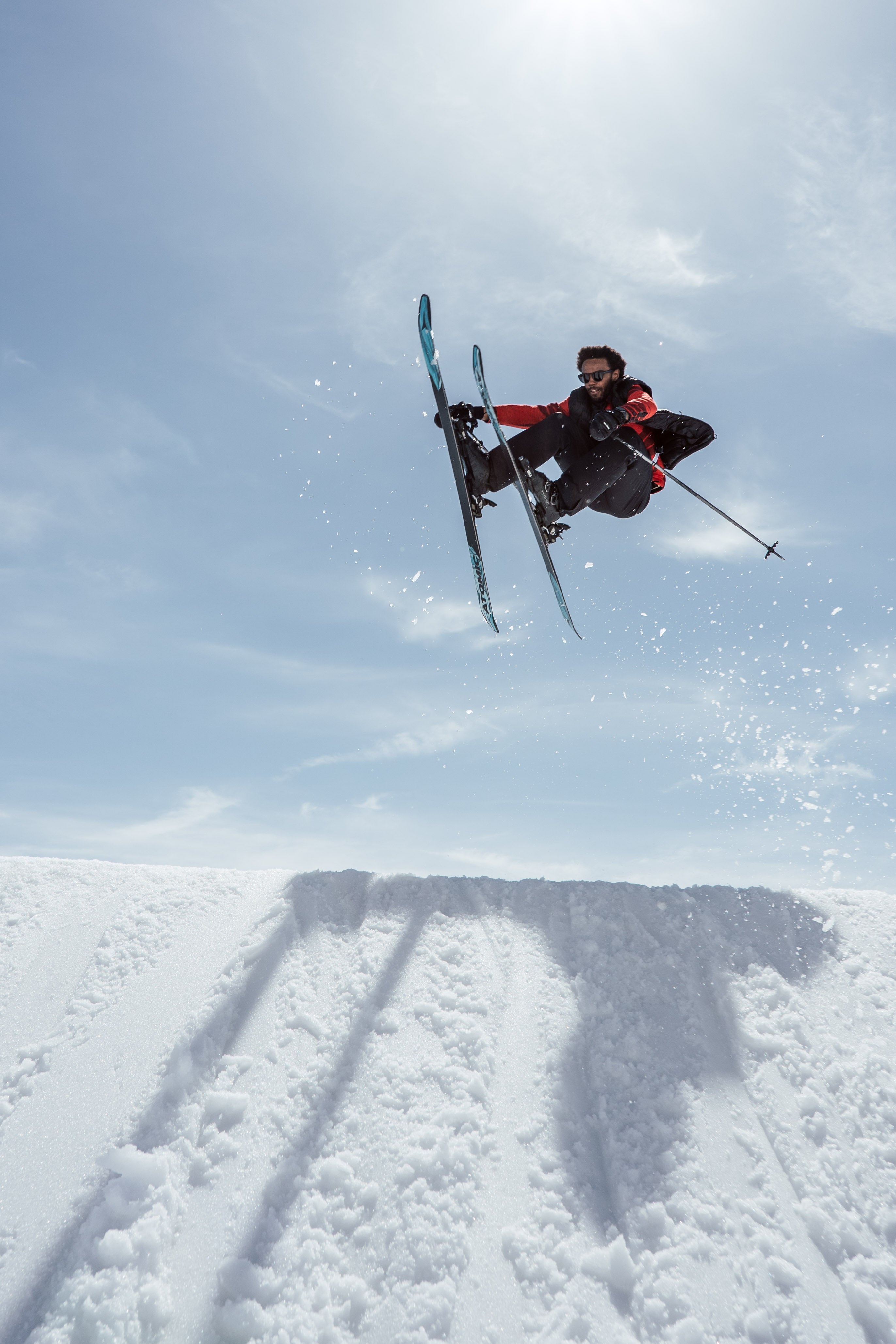 Dennis Renalter mid-air performing a jump over a snowy ramp under a bright blue sky, wearing a black and red ski outfit with poles.