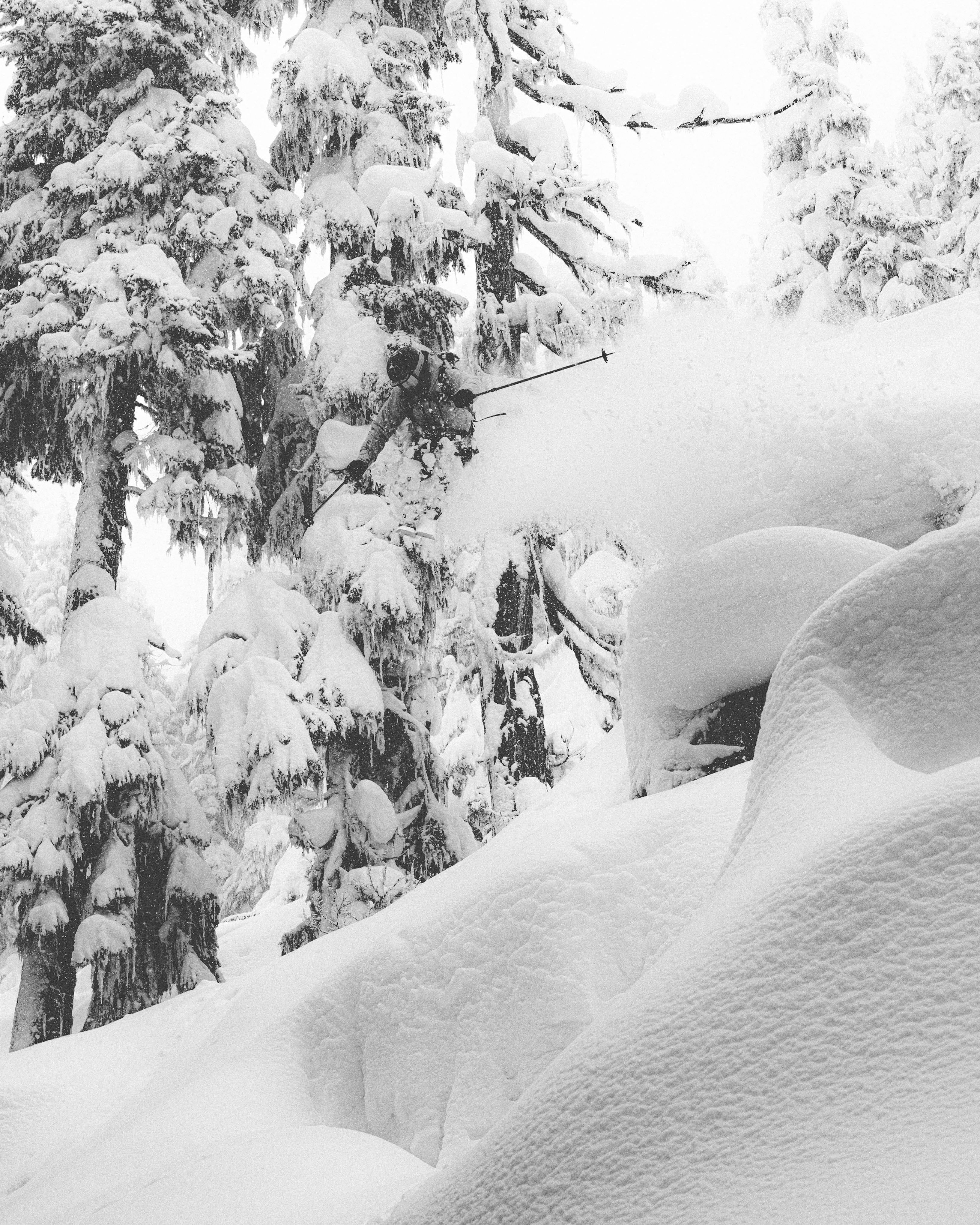Crhistopher Rubnes dropping in the middle of a snow-covered forest with thick drifts of snow in the foreground and tall pine trees heavily laden with snow in the background.