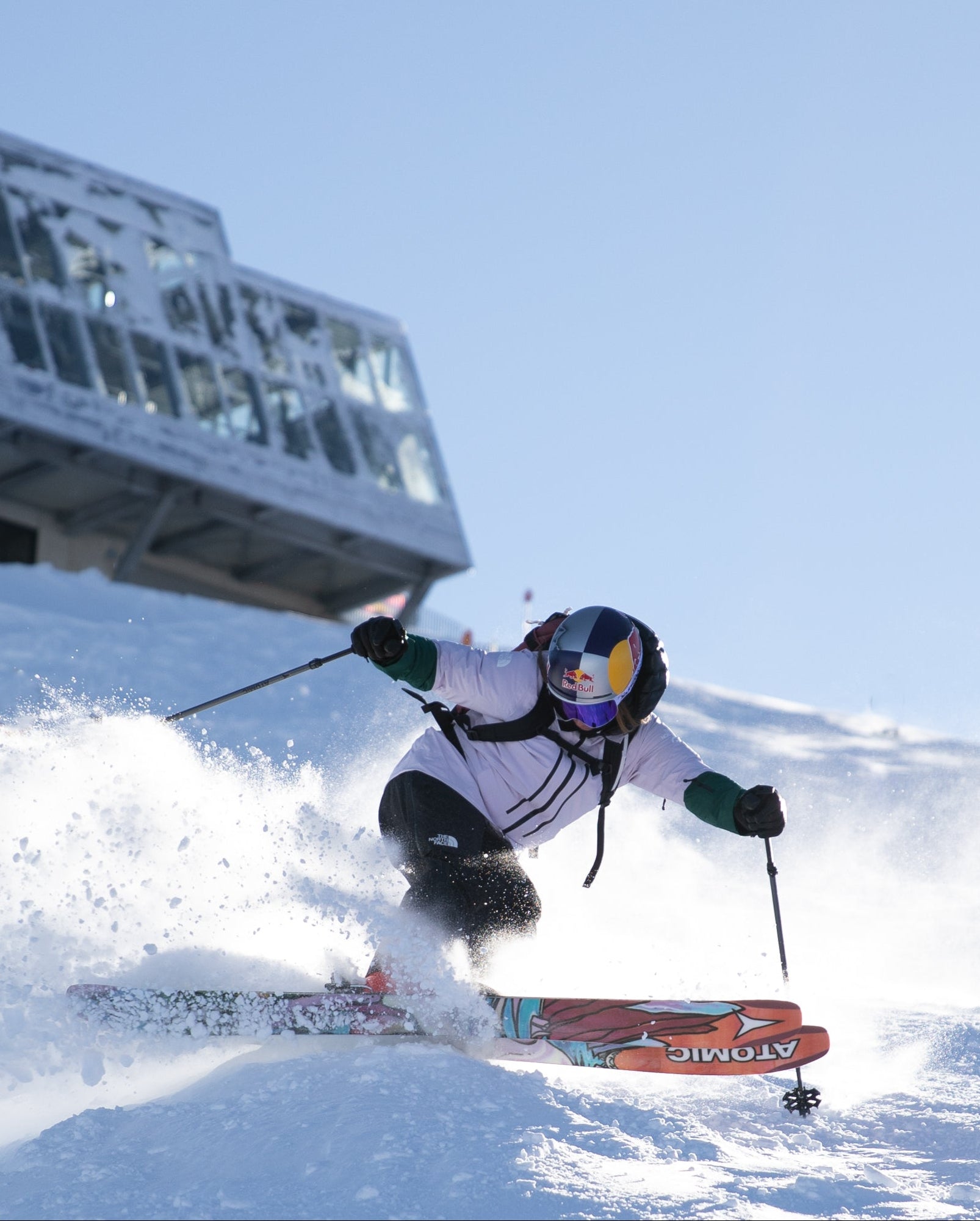 Arianna Tricomi carving through fresh powder on a snowy slope, wearing a white ski jacket, black pants, and a helmet with a blue visor, with a ski lift and modern building visible in the background under a clear blue sky.