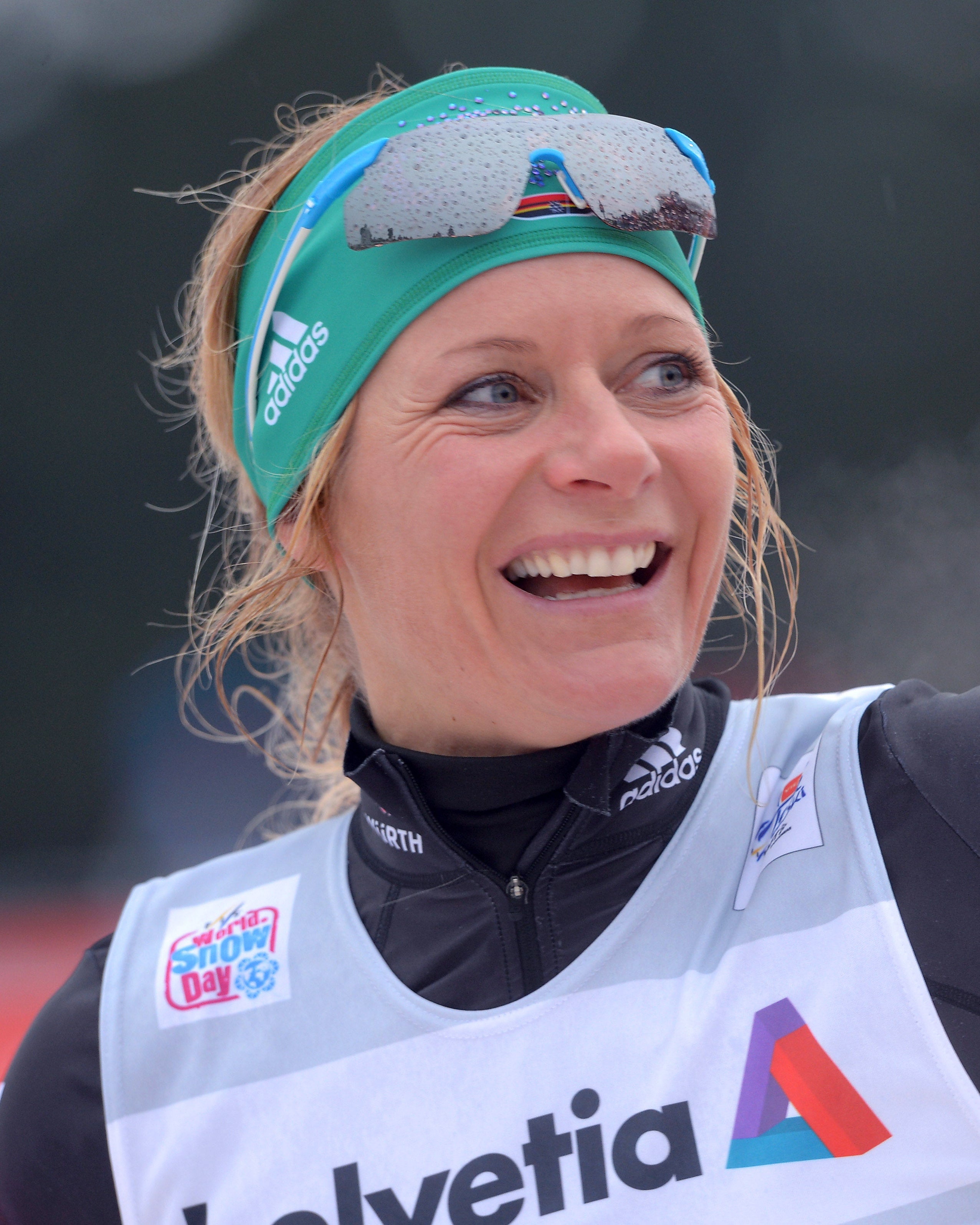 Claudia Nystad smiling and raising her arm, wearing a green Adidas headband with ski goggles, a black and red long-sleeve jersey, and a white bib with the word 'Helvetia' and a colorful triangular logo.