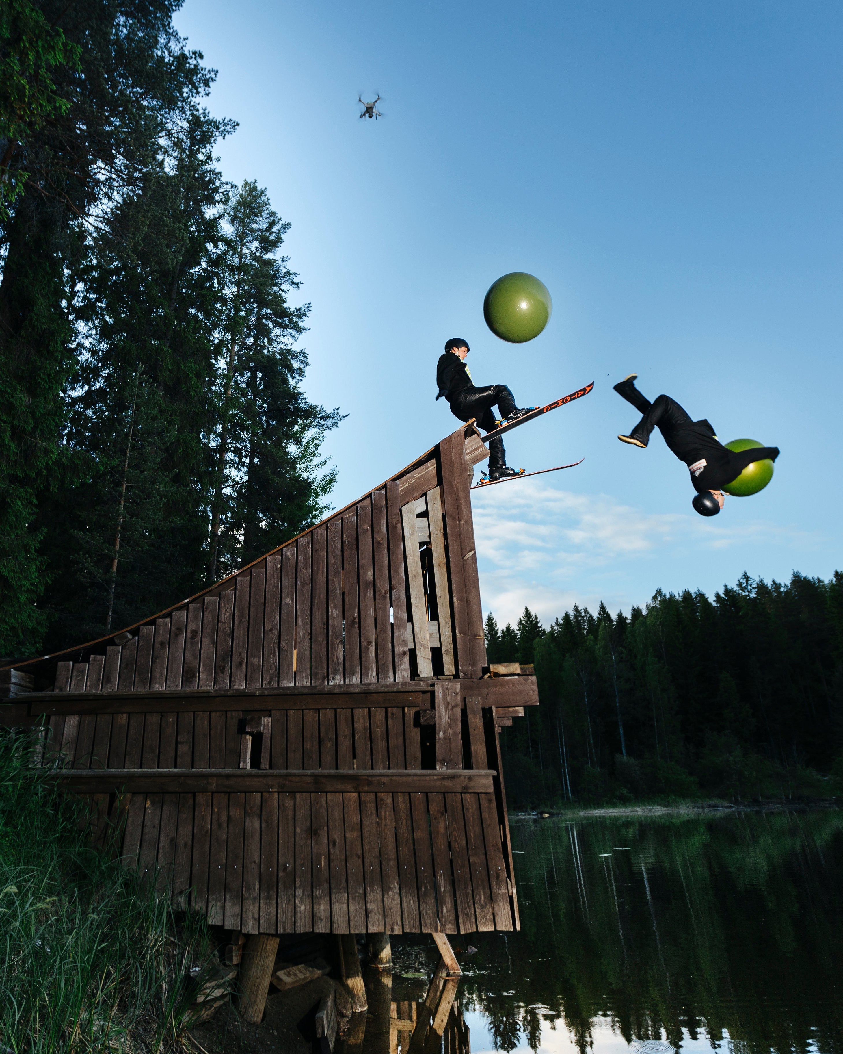 Two people performing a high-trick tandem rope jump on a wooden platform over a lake, with a large green ball in the air between them under a clear blue sky and forest background.