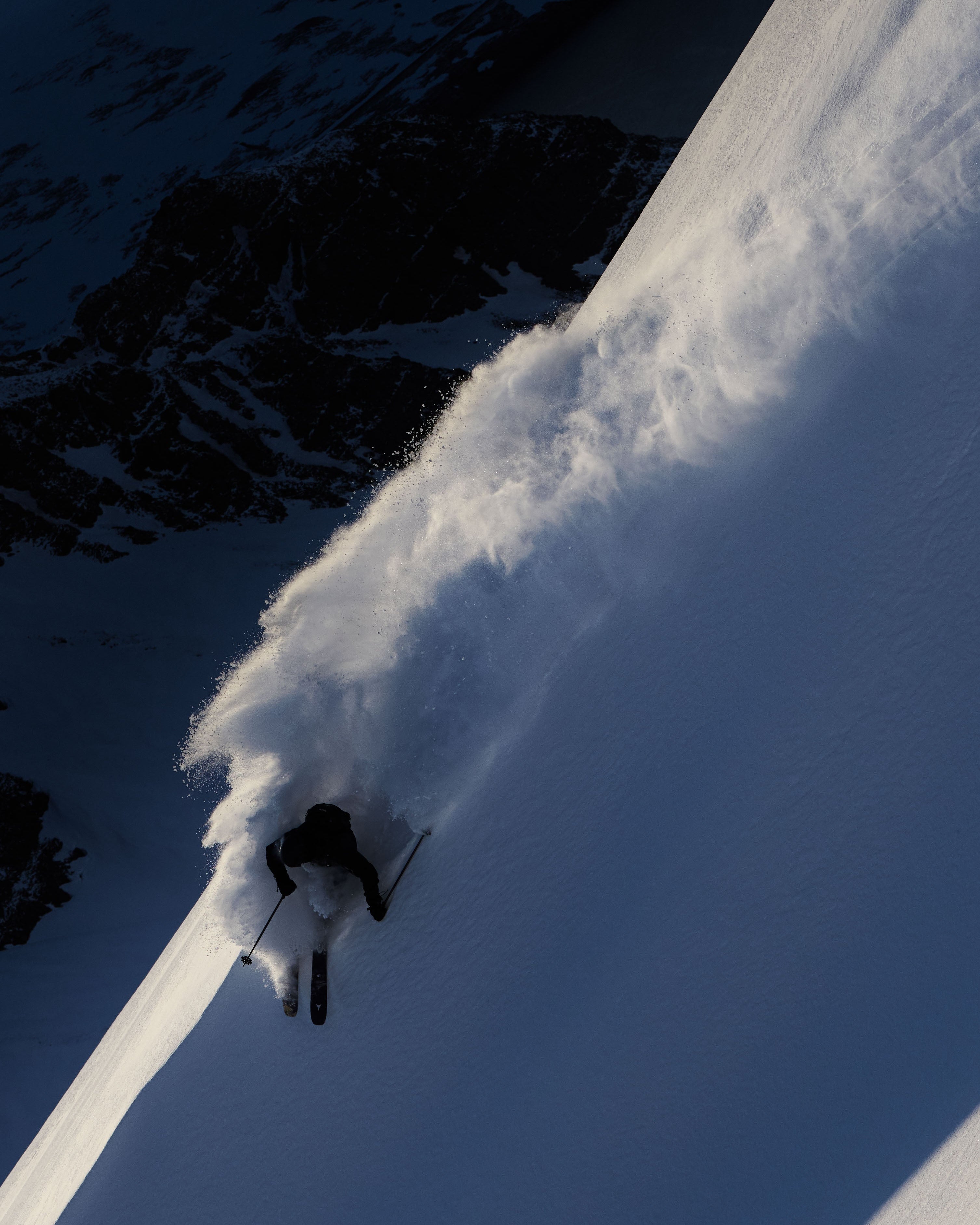 Chris Benchetler carving through deep powder snow on a steep mountain slope, with dark rocky terrain visible in the shadowed upper left.