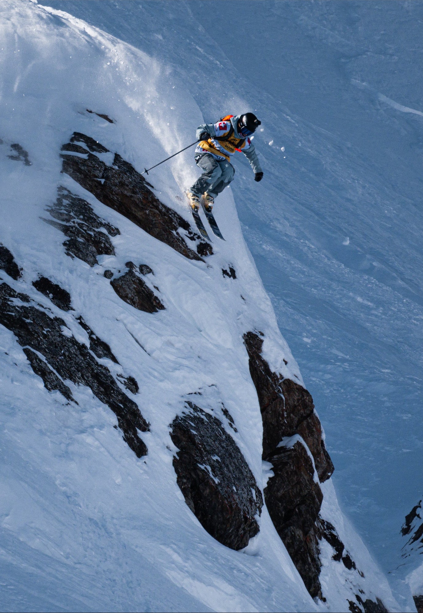 Sybille Blanjean in a blue and orange outfit navigates a steep, snow-covered mountain slope with exposed dark rock, captured from an elevated angle.