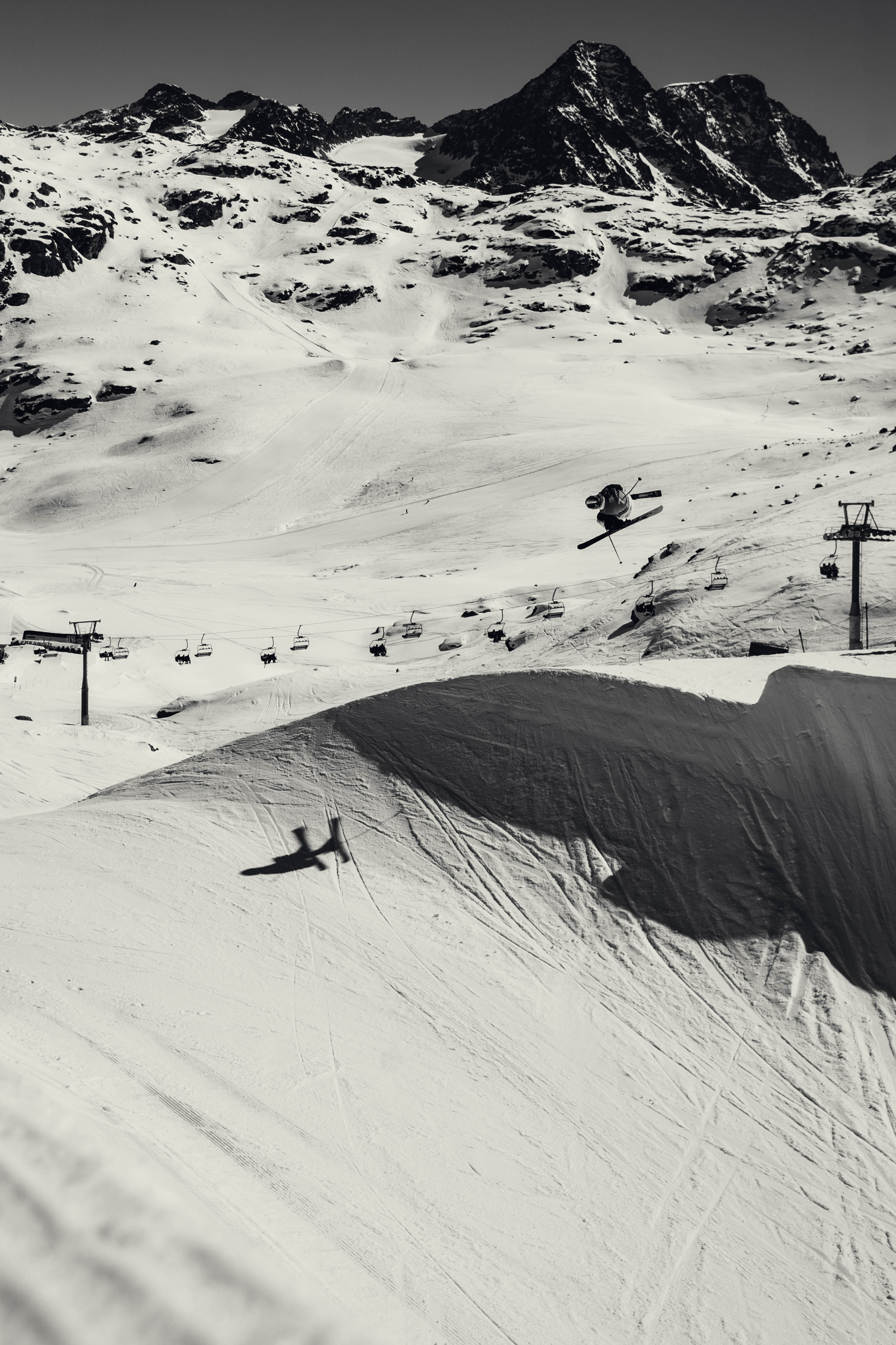Black and white photograph of a snowy mountain landscape with ski lifts, Fabian Boesch in midair, and a ski jump slope in the foreground.