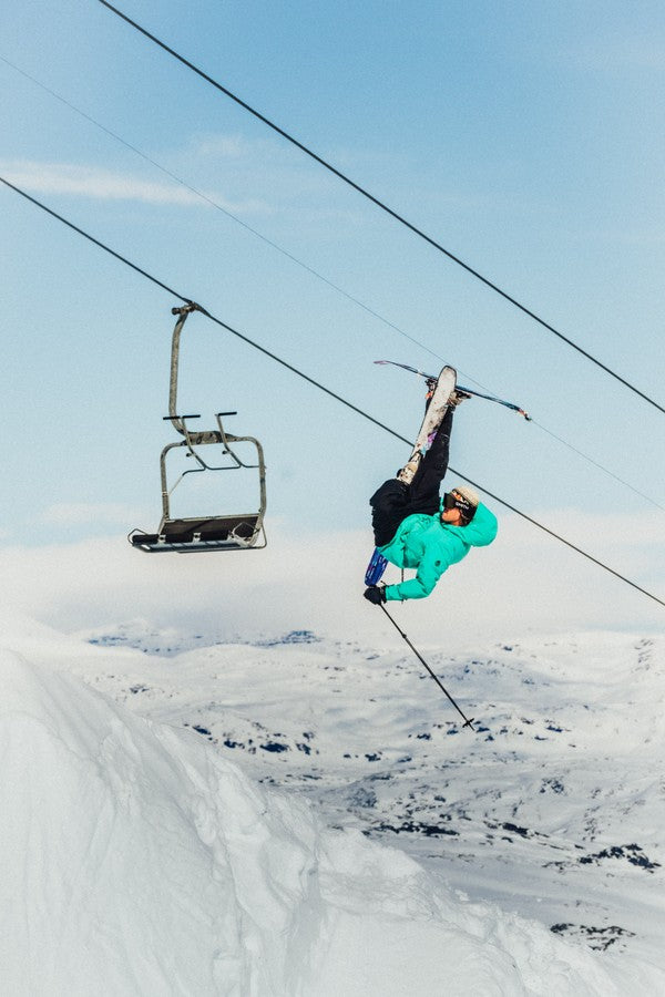 Bobby Brown performs a backflip trick in midair on a snowy mountain slope, with a chairlift and blue sky in the background.