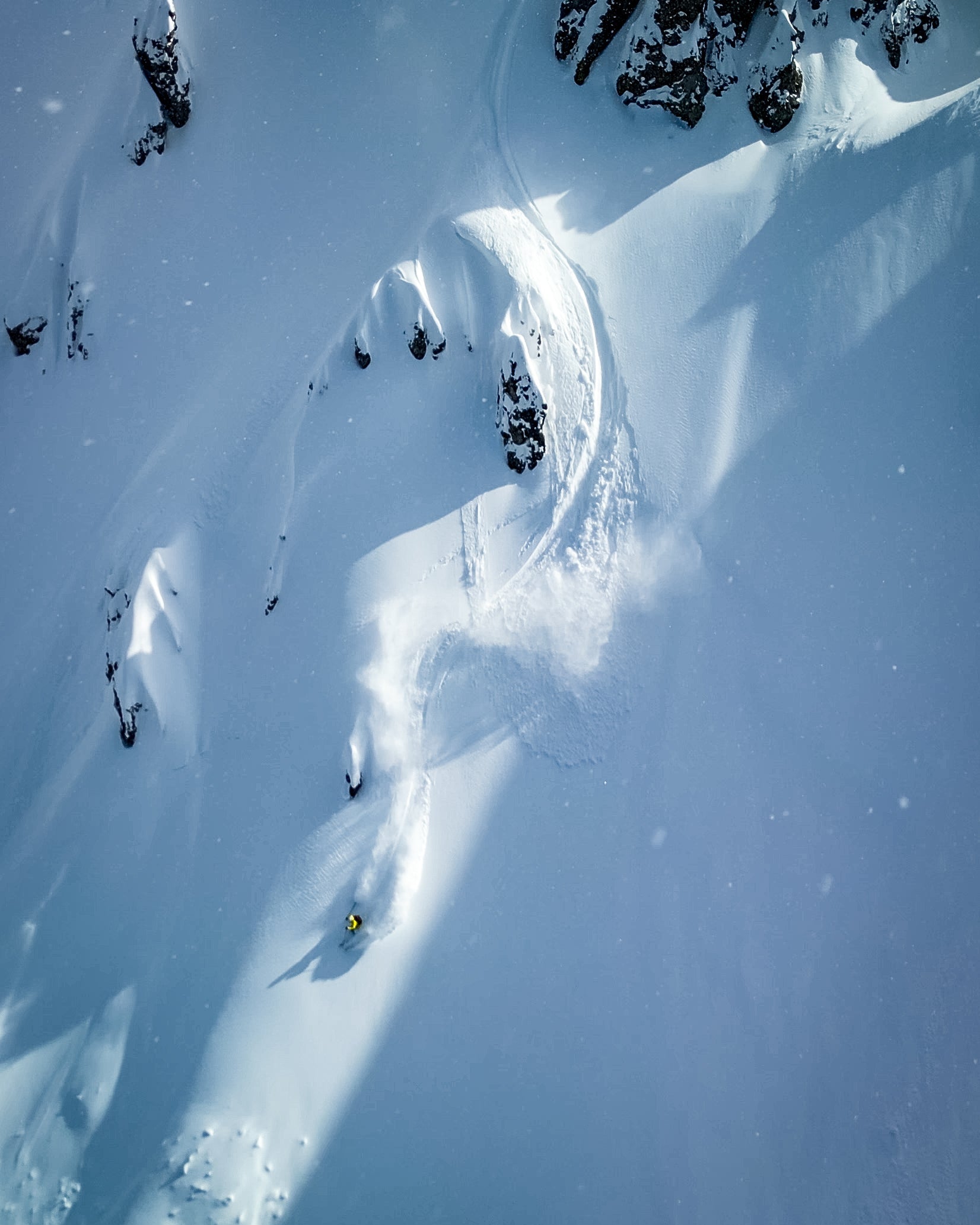 Aerial capture of Amie Engerbretson descending in a snowy mountain slope with scattered dark rock outcrops.
