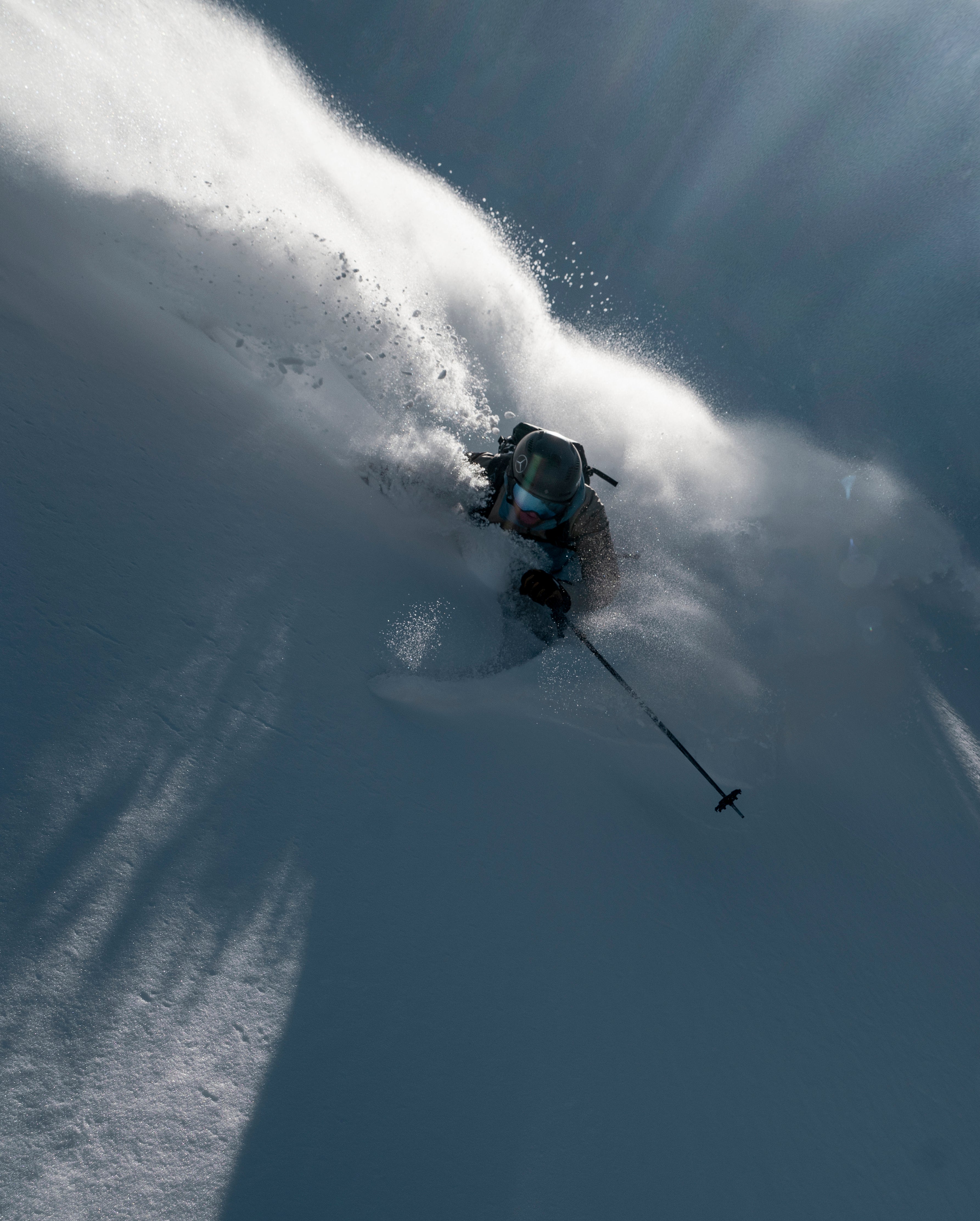 Matthias Haunholder carving through deep powder snow, creating a dramatic spray of white snow and casting long shadows under bright sunbeams.