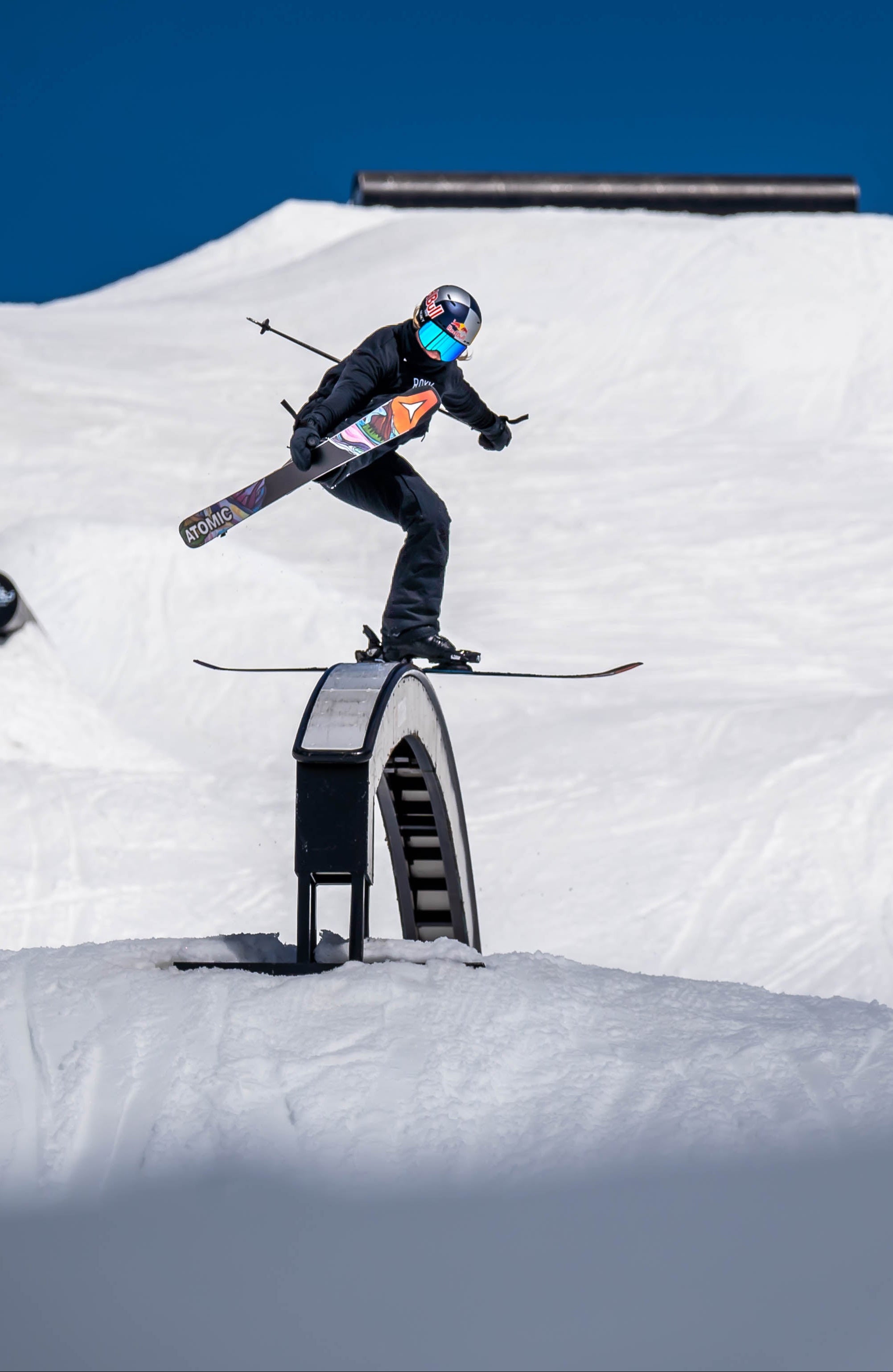 Tess Ledeux in black gear and a blue helmet jumps over a small metal rail on a snowy mountain slope under a clear blue sky.