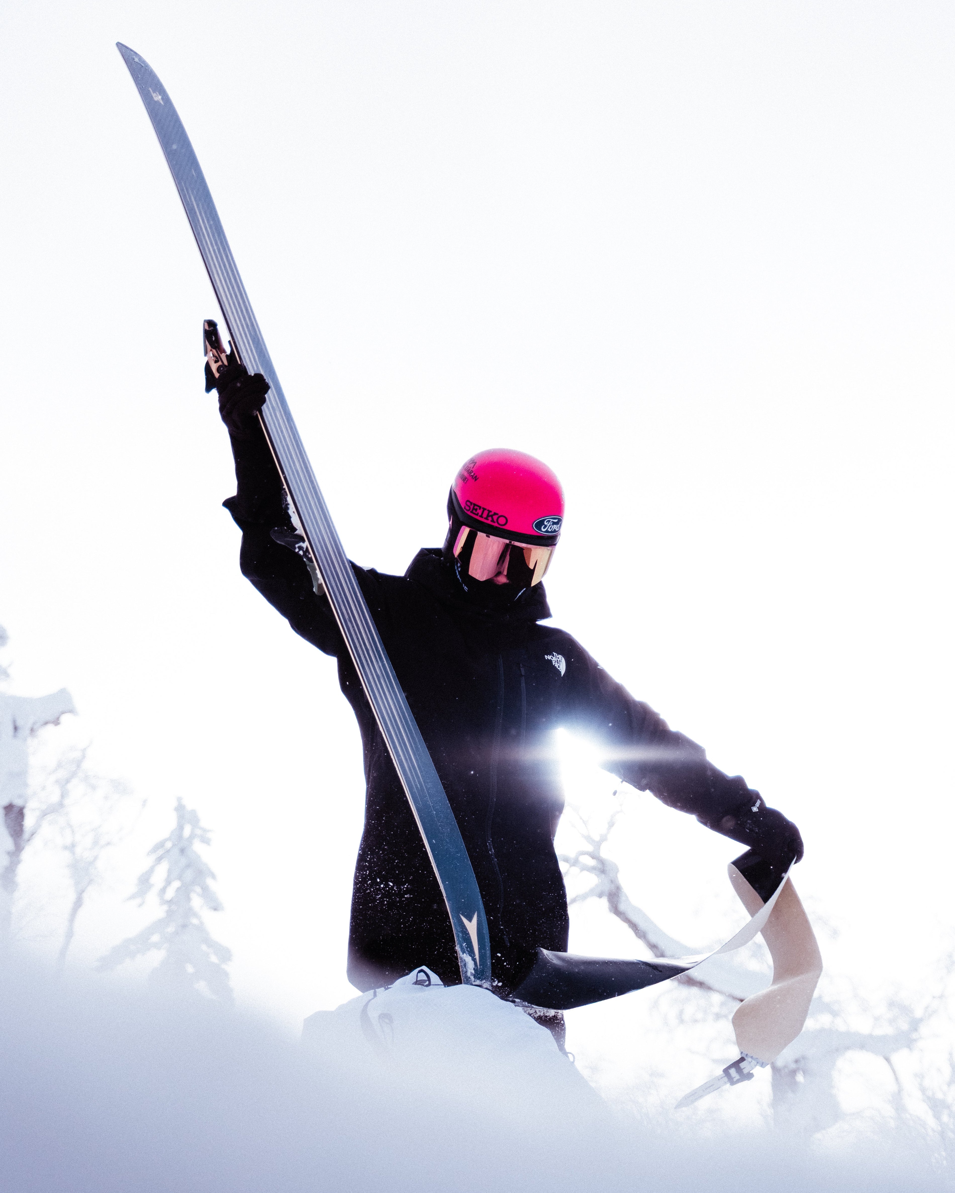 Aymar Navarro in a black ski jacket and pants, wearing a bright pink helmet and reflective goggles, holds up a ski against a bright, snowy outdoor background.
