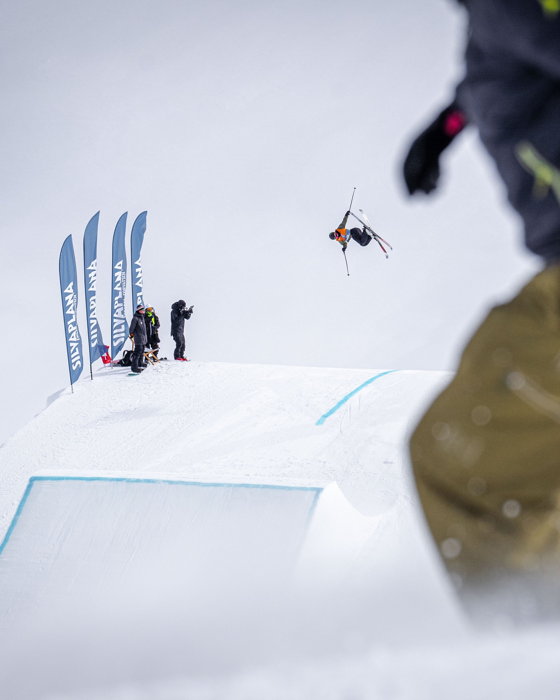 A snowy outdoor scene with Megan Oldham mid-air performing a jump in the distance, with a blurred foreground figure wearing dark clothing and olive pants. Snowboarders and flags are visible on the snowy ramp in the background.