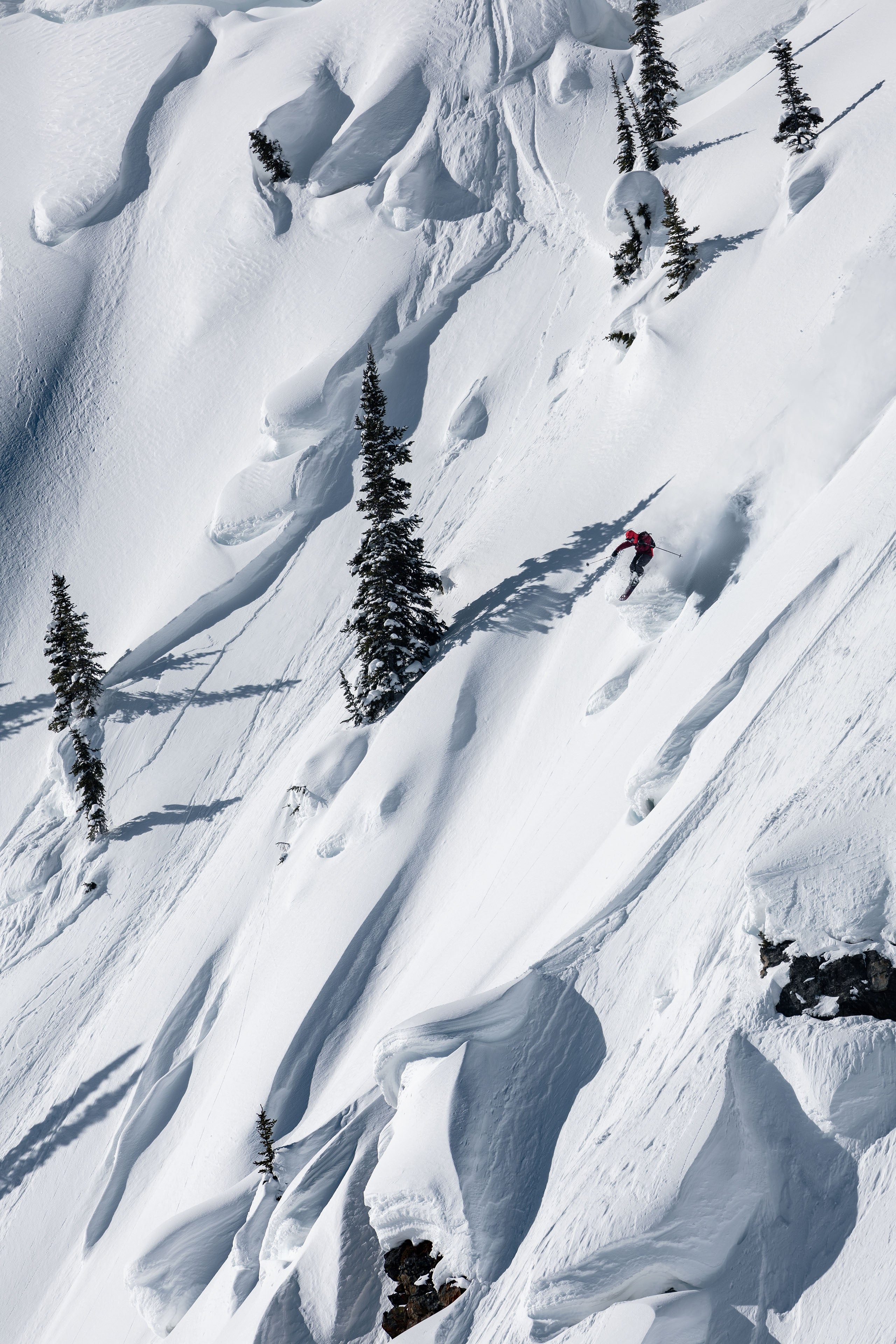 Aerial view of Dennis Renalter carving through deep powder snow on a steep mountain slope, with scattered evergreen trees casting long shadows.