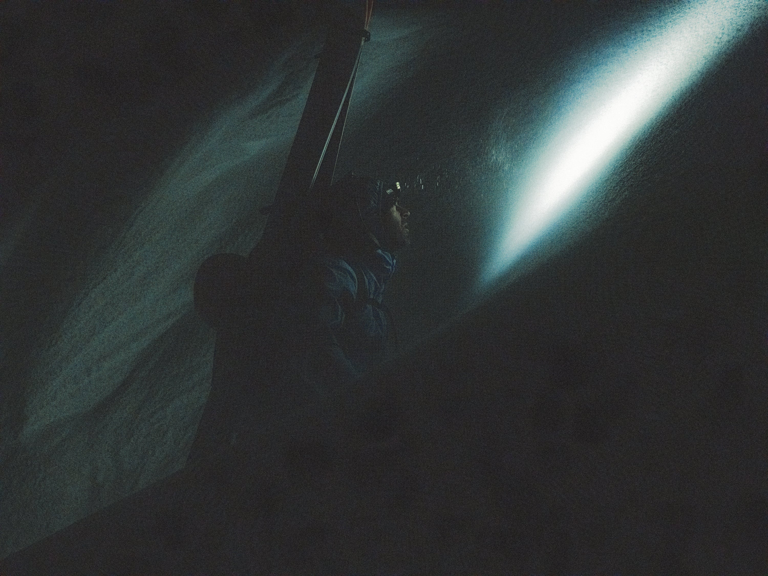 Chirstopher Rubens in a dark, low-light environment wearing a headlamp that casts a bright beam of light across a snowy or rocky surface.