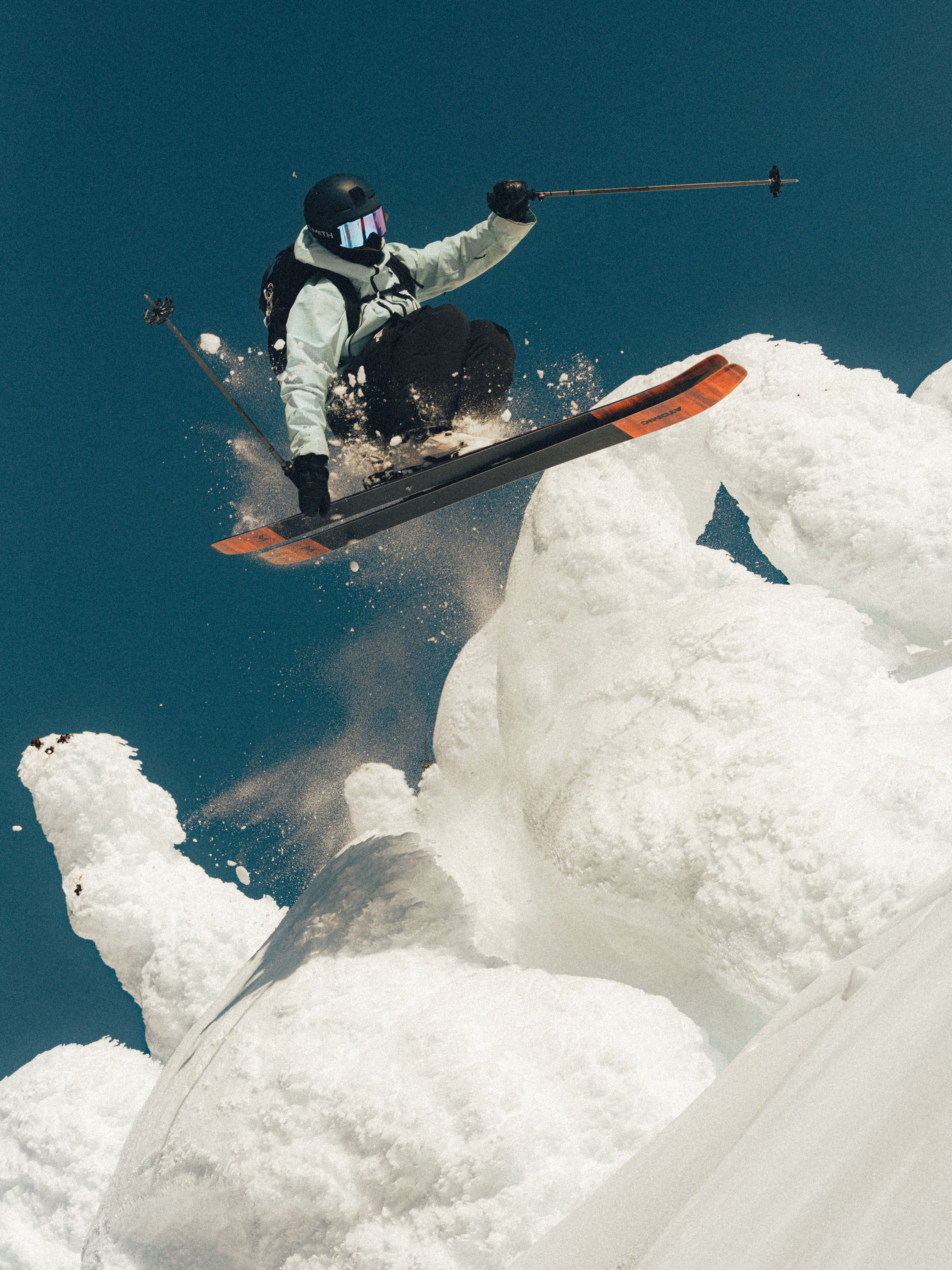 Janelle Yip above a snowy ridge, wearing a black helmet, white jacket, and black pants, with orange ski tips visible against a deep blue sky.