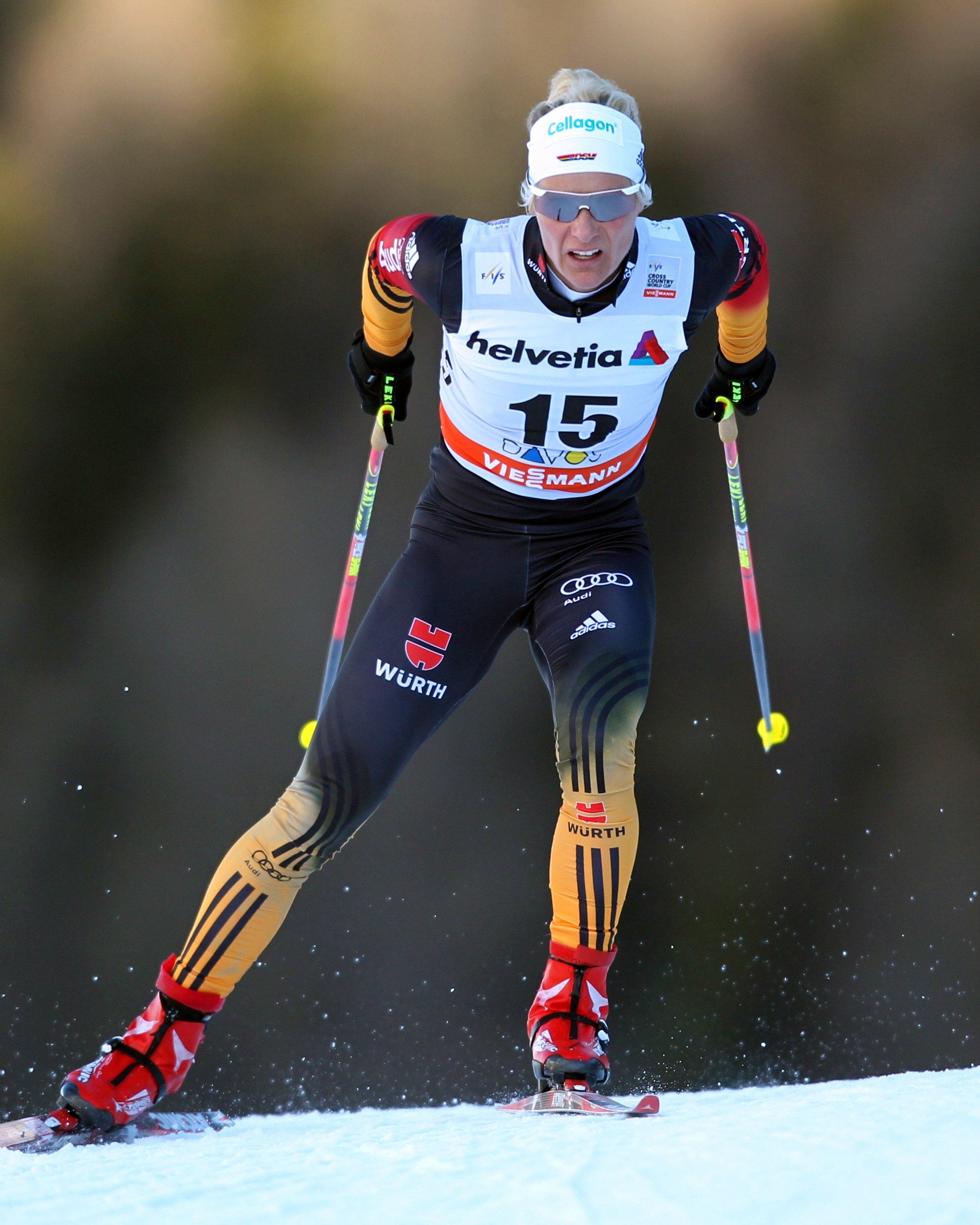 Claudia Nystad in a black, white, and yellow racing suit with bib number 15, skiing through the snow on a sunny day.