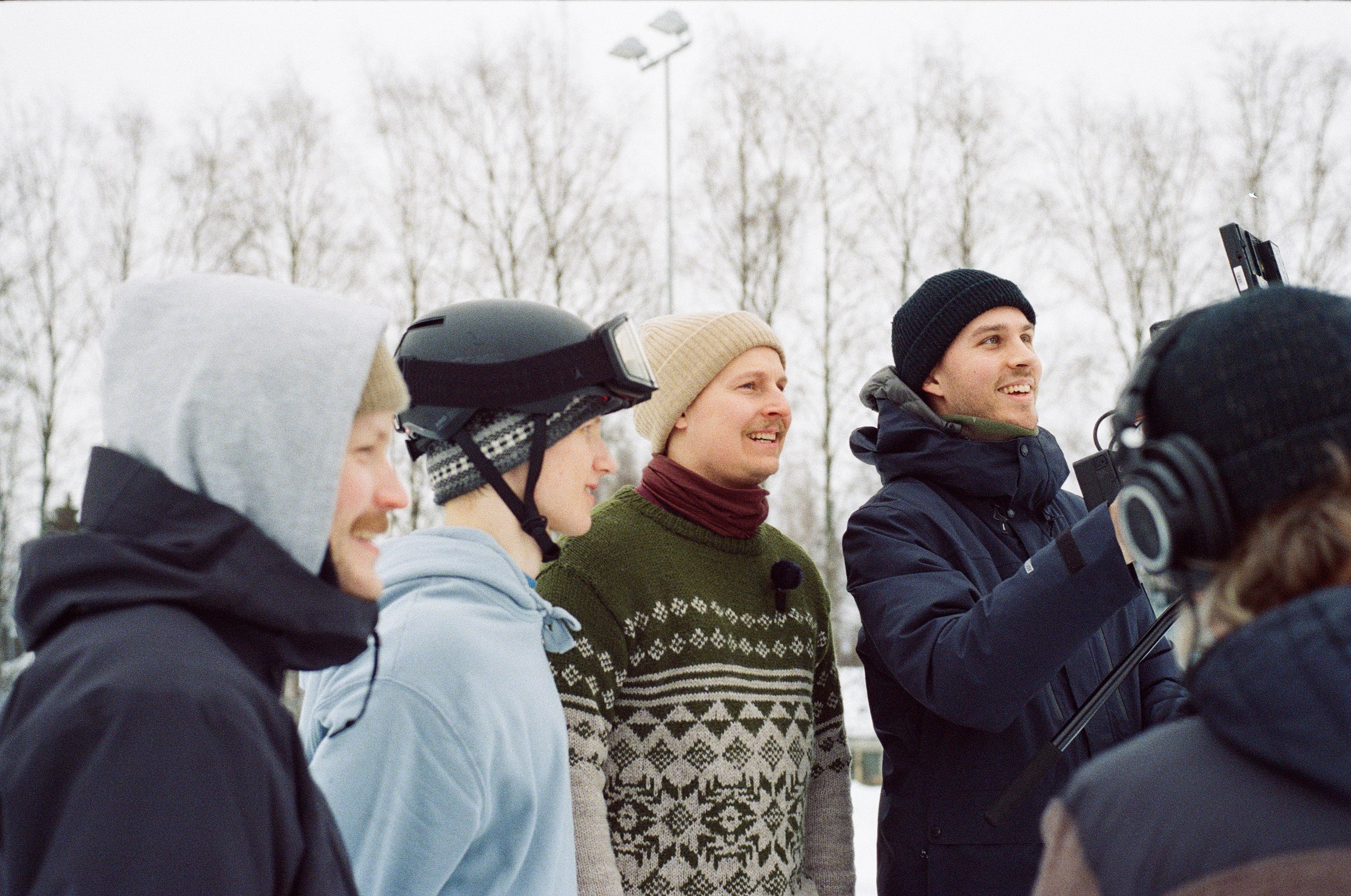Real skifi in winter clothing, including beanies, a patterned knit sweater, and jackets, with one holding a camera. Bare trees and an overcast sky are in the background.