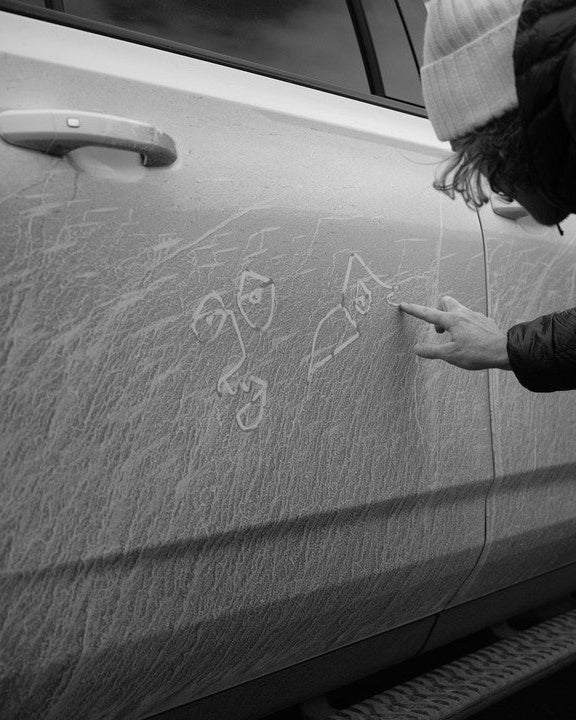 Chris Benchetler wearing a white beanie, sketching his signature artwork onto a snow-dusted car door in black and white.